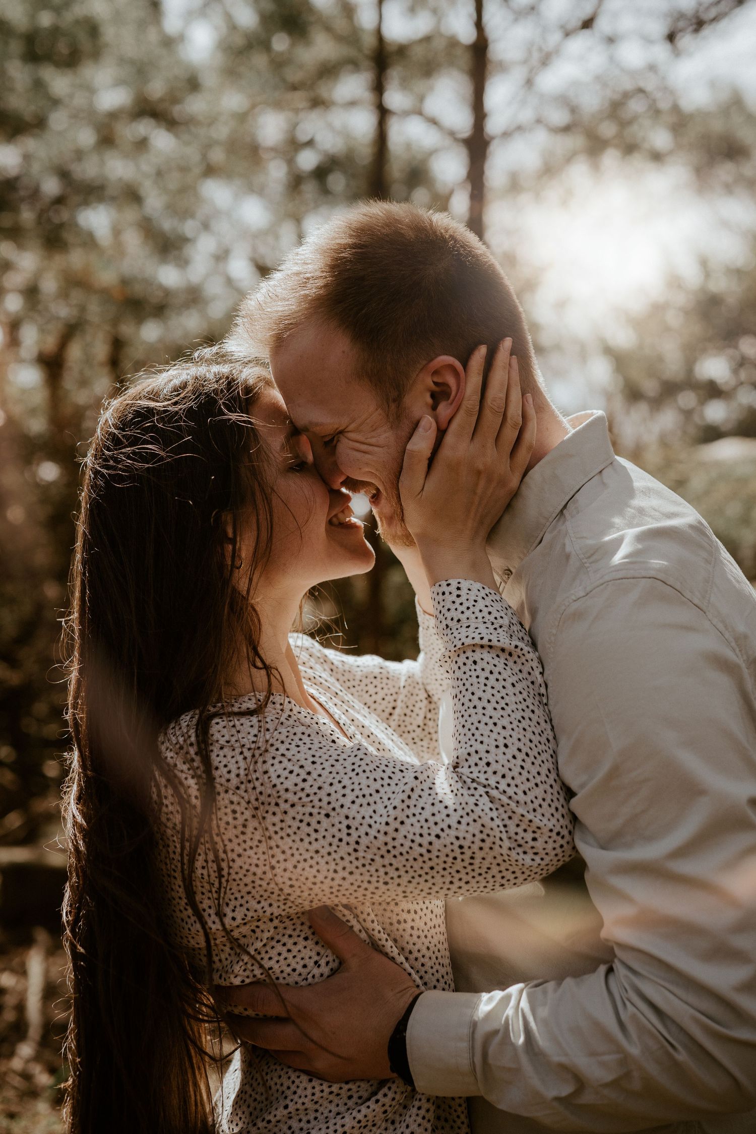 Intimate romantic moment captured in sunlit forest as couple in white embraces during outdoor photoshoot.