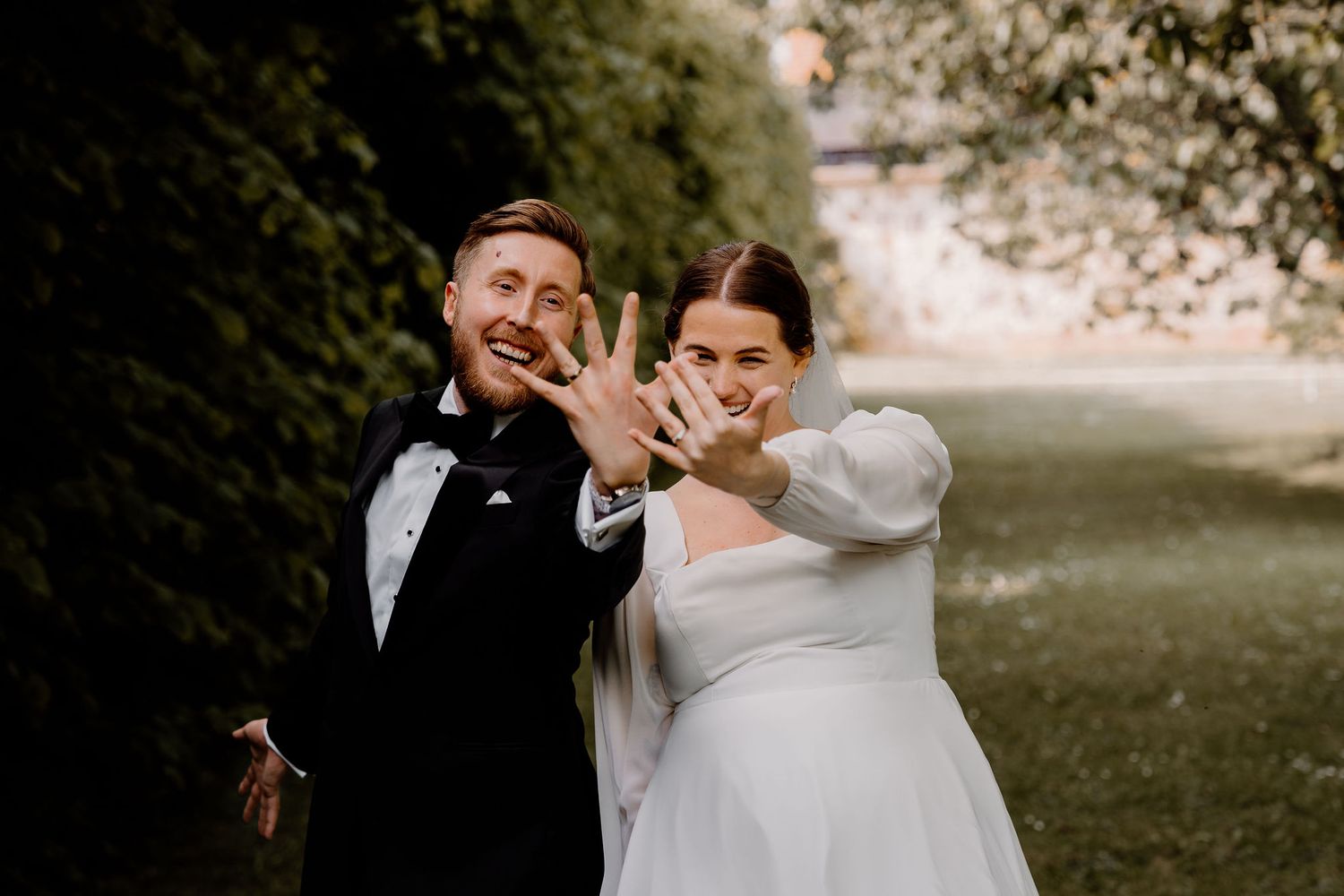 A happy couple in wedding attire show off their rings while posing together outdoors in a natural setting.