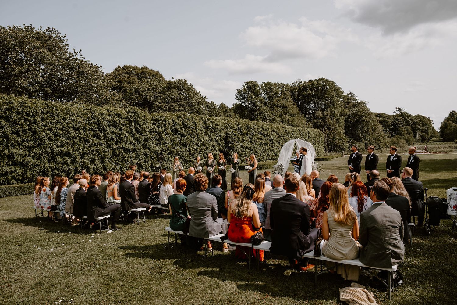 An outdoor wedding ceremony on a grassy lawn with guests seated in white chairs facing an altar under cloudy skies.