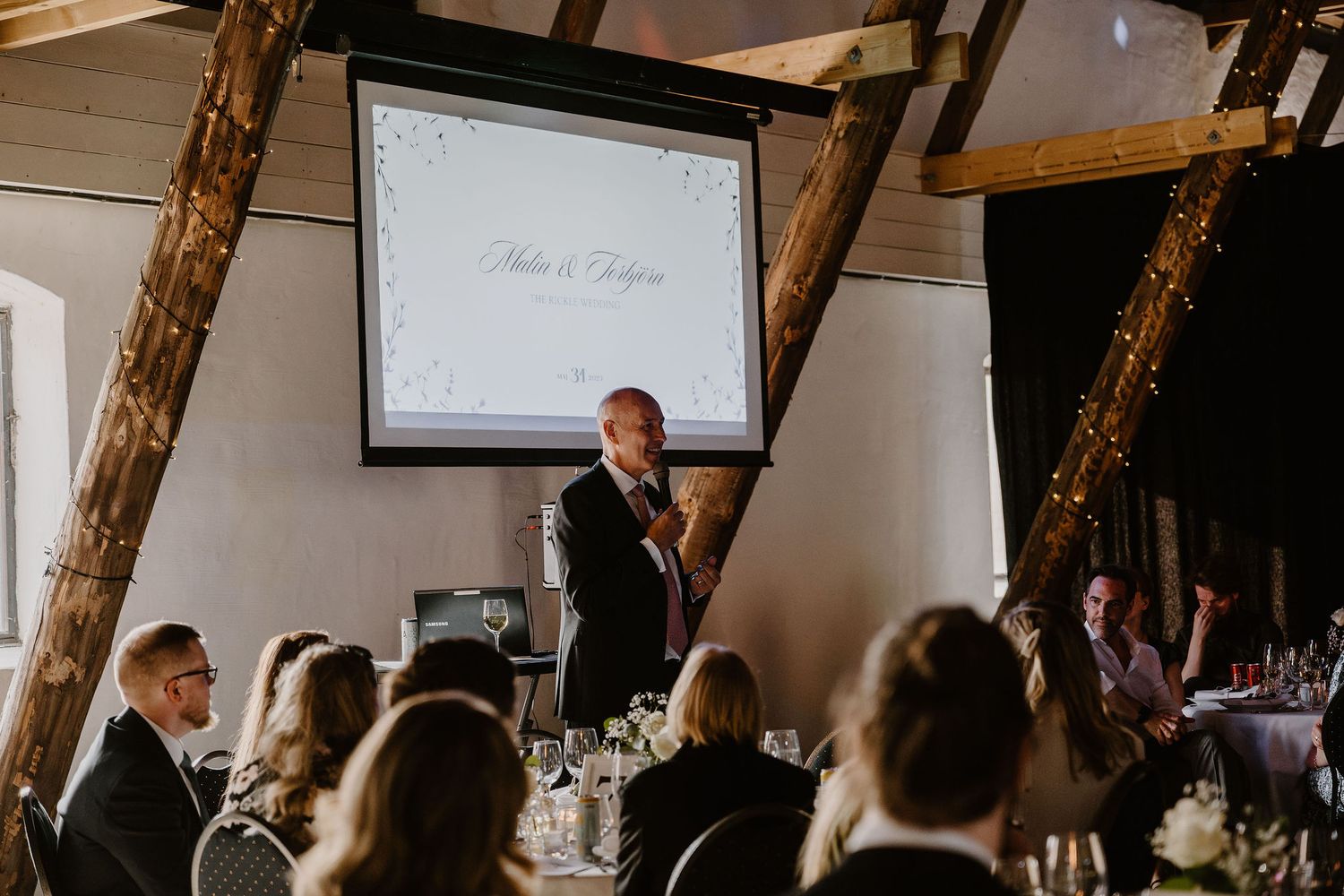 Wedding guests listen attentively as a speaker gives a toast in a rustic venue with exposed wooden beams and a projector screen.