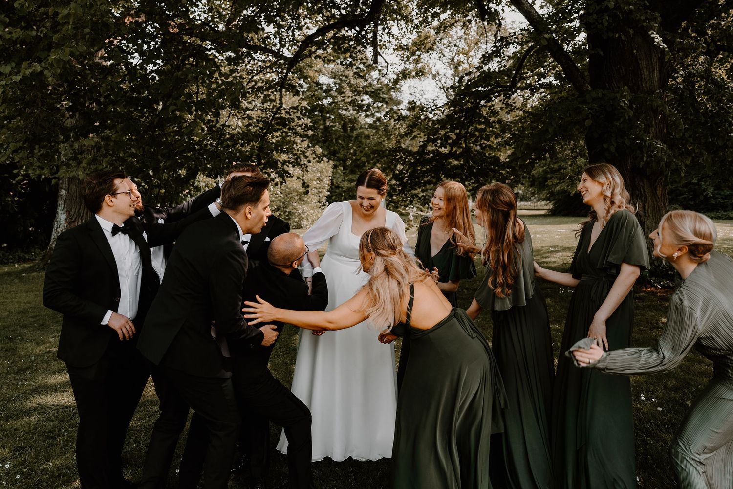 Wedding party gathered outdoors in formal attire with bridesmaids in dark green dresses and groomsmen in black suits.