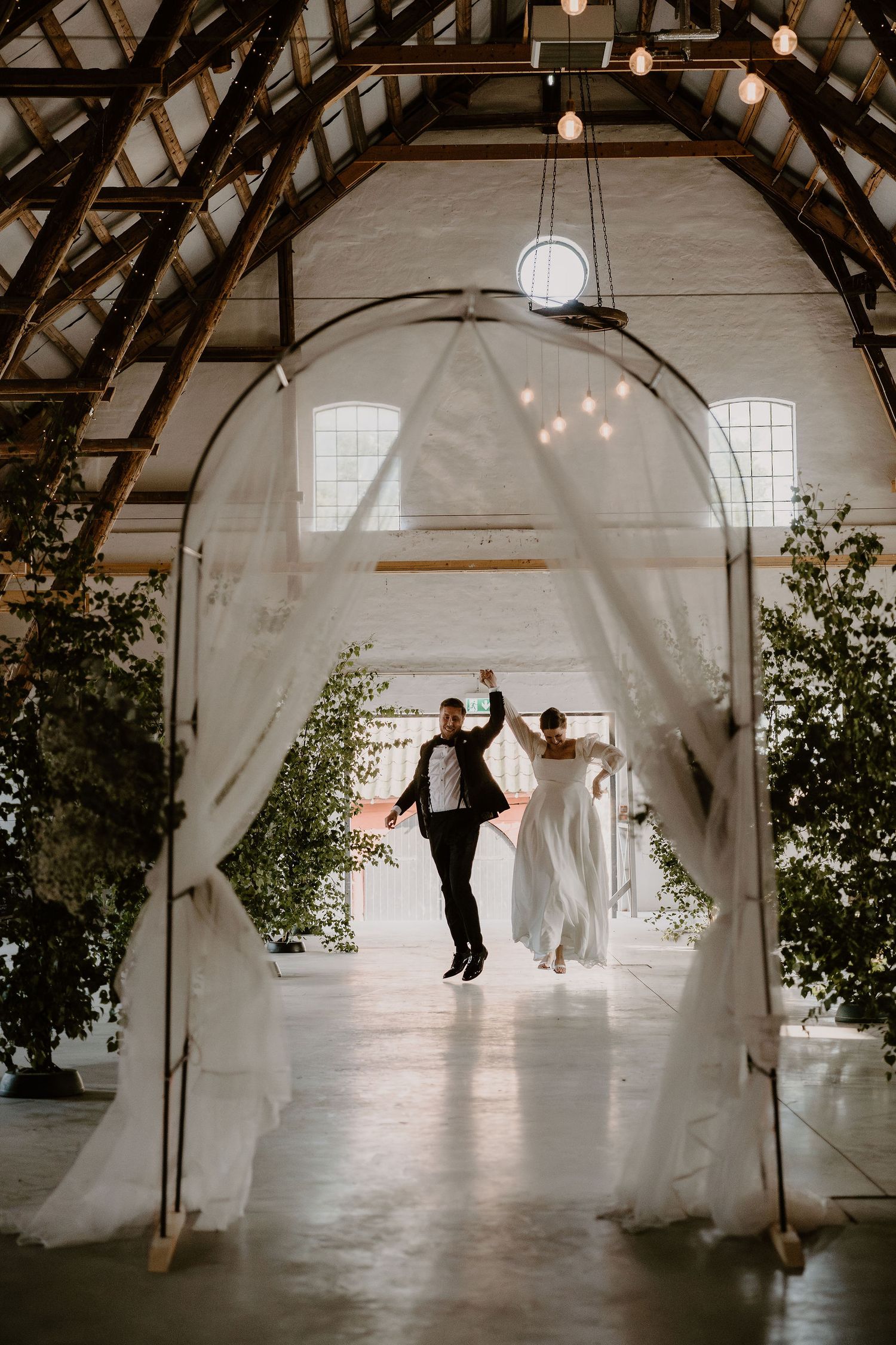 Under a white draped archway with string lights in a barn-like venue, a couple celebrating joyfully on their wedding day.