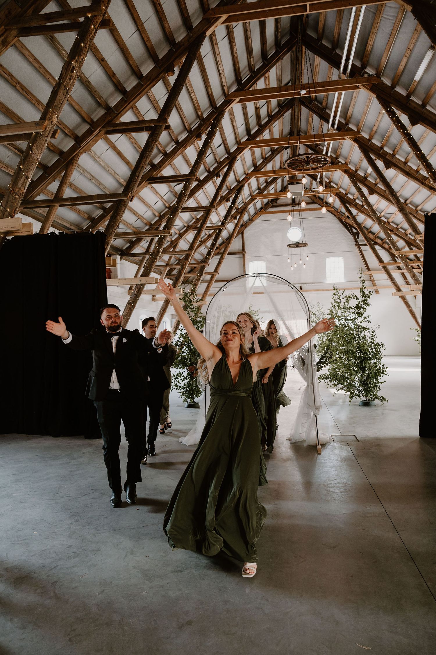 Wedding party members walk through a rustic barn venue with exposed wooden beams and white walls.