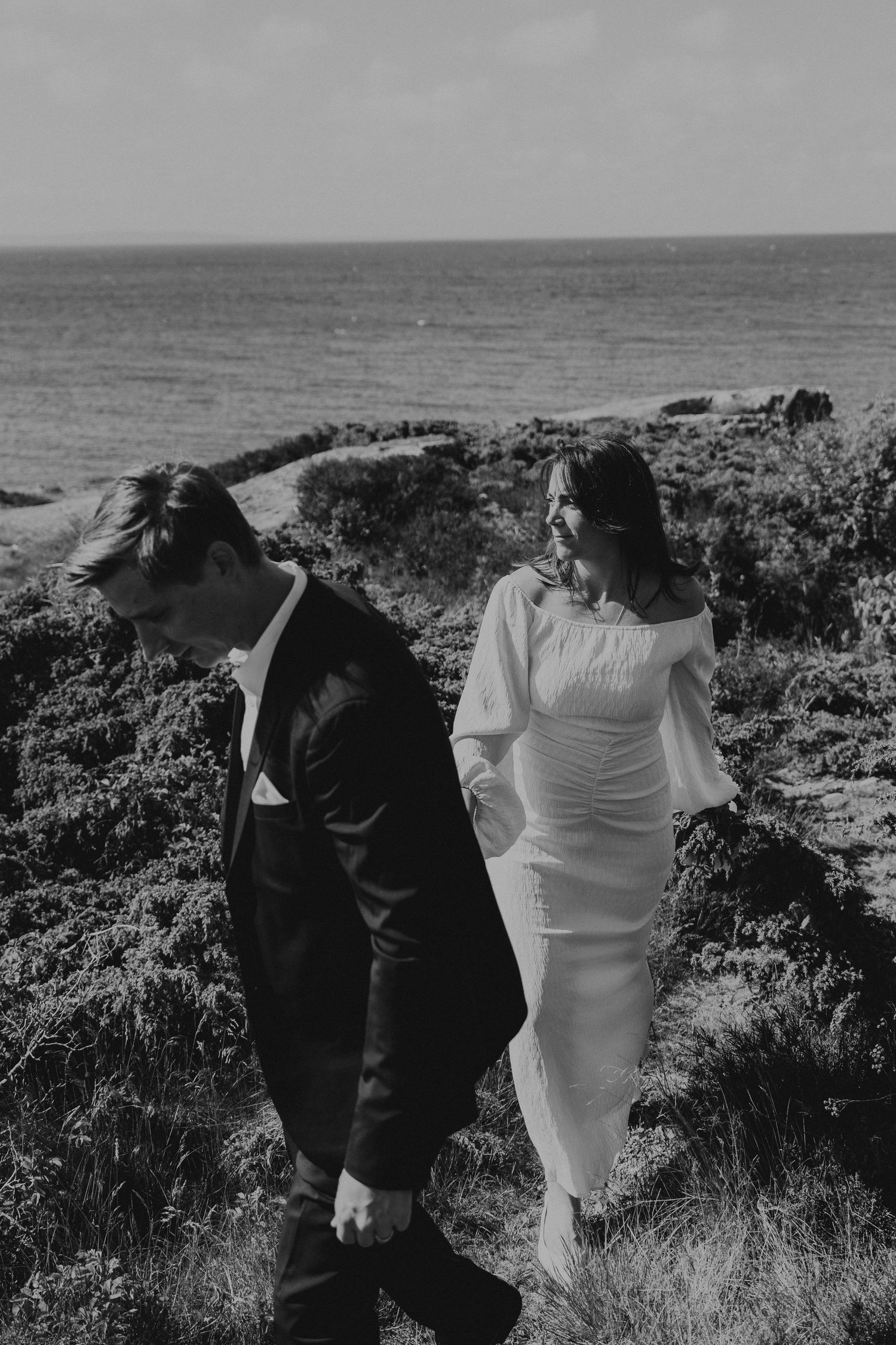 A couple in formal attire walks away from the camera along a coastal cliff path overlooking the ocean.