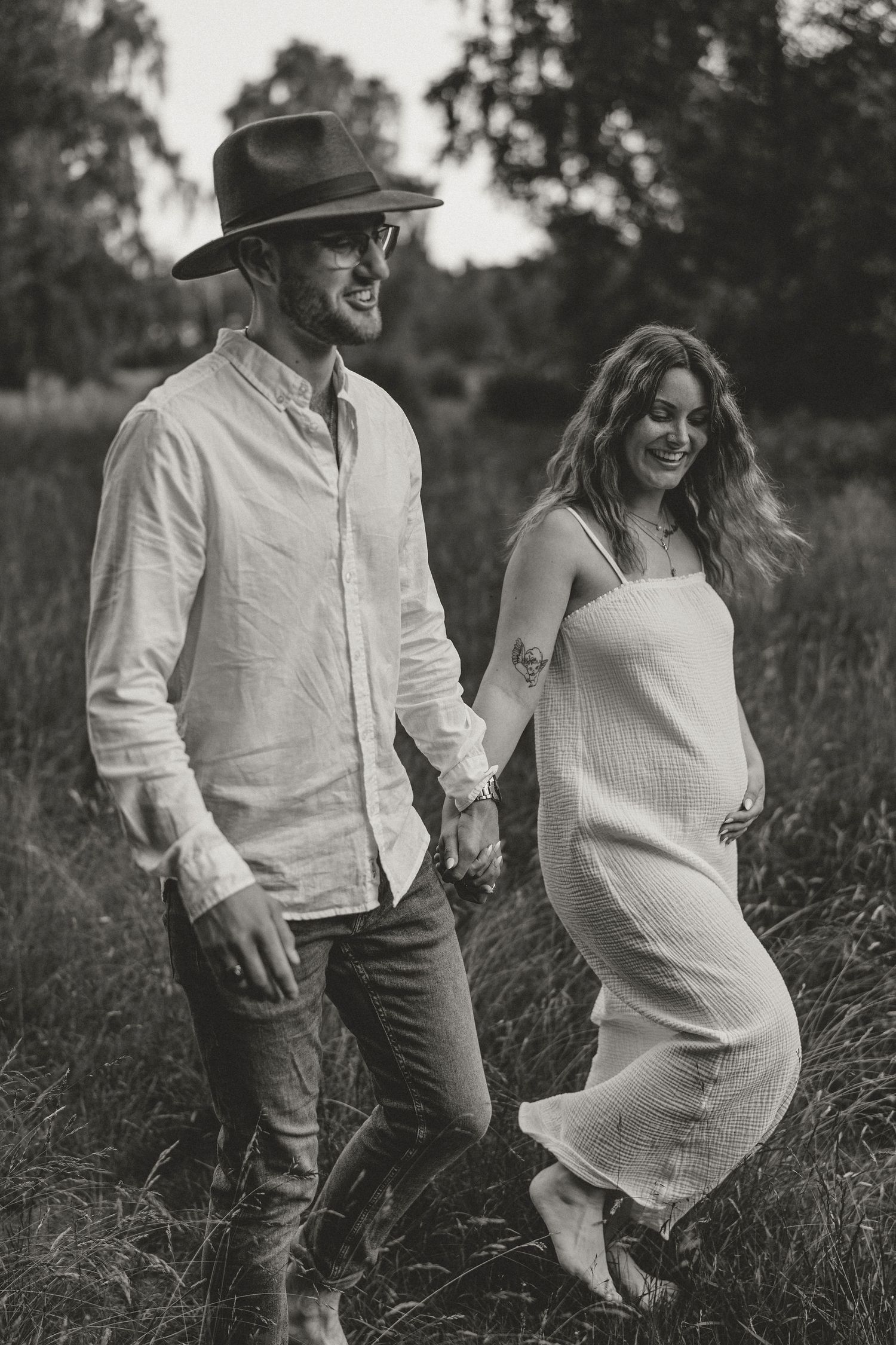 Black and white photo of a couple walking together in a field, with man in hat and woman in flowing dress.