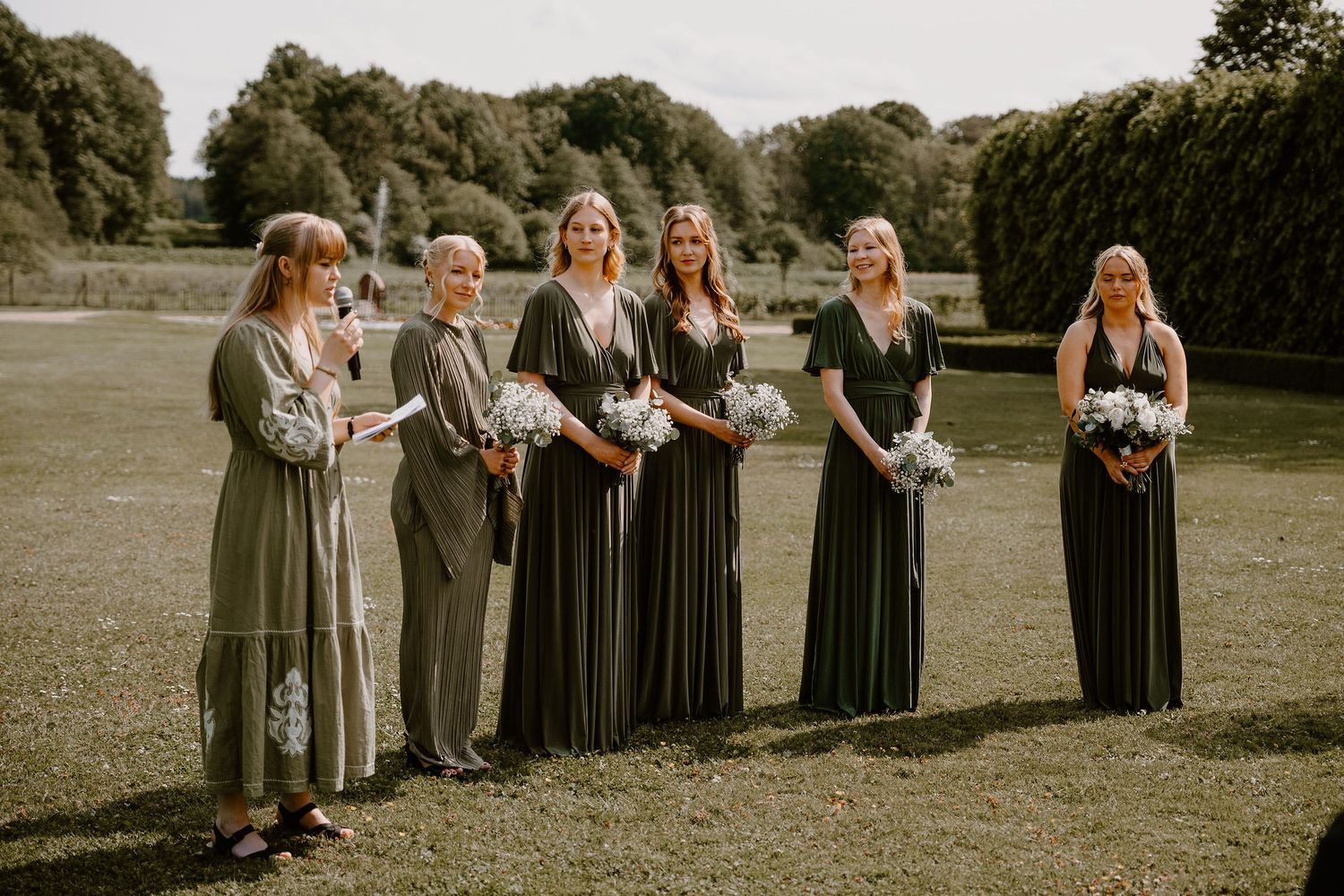 A line of bridesmaids in gray dresses standing on a gravel path during an outdoor wedding ceremony.