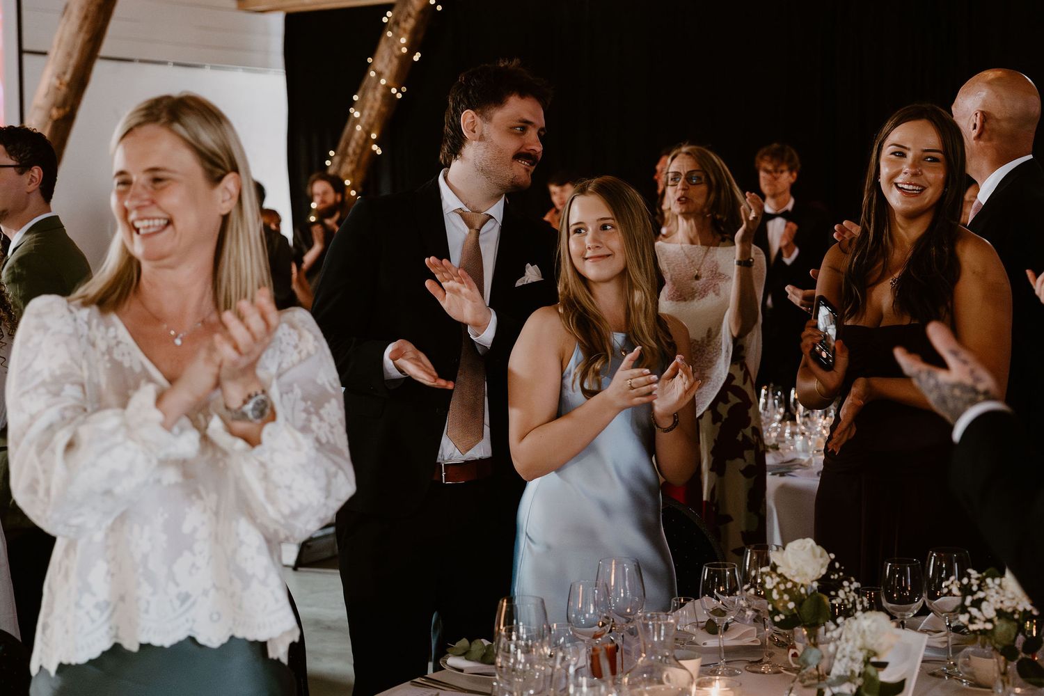Joyful wedding guests seated at elegant reception tables raise their glasses in a celebratory toast.