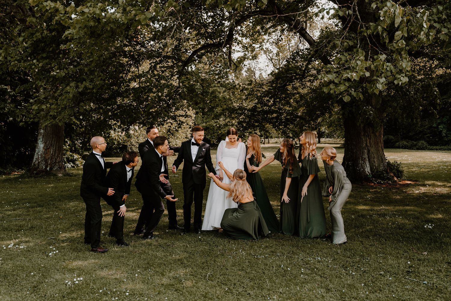 The wedding party poses together outdoors during a formal portrait session beneath large trees on a sunny day.