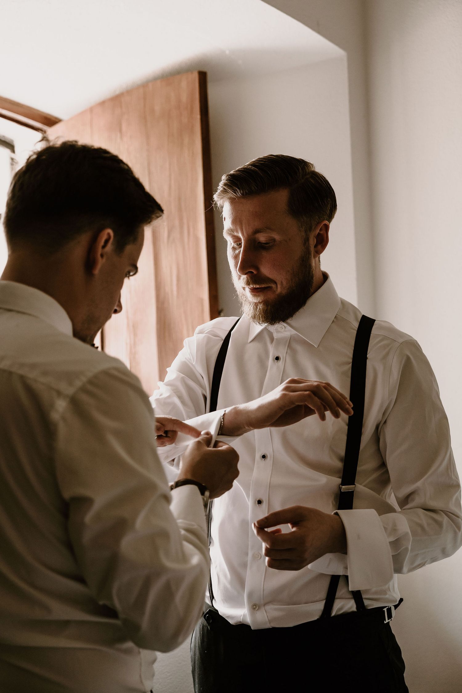 Two people in white dress shirts and suspenders help each other with formal attire preparations in a bright room.
