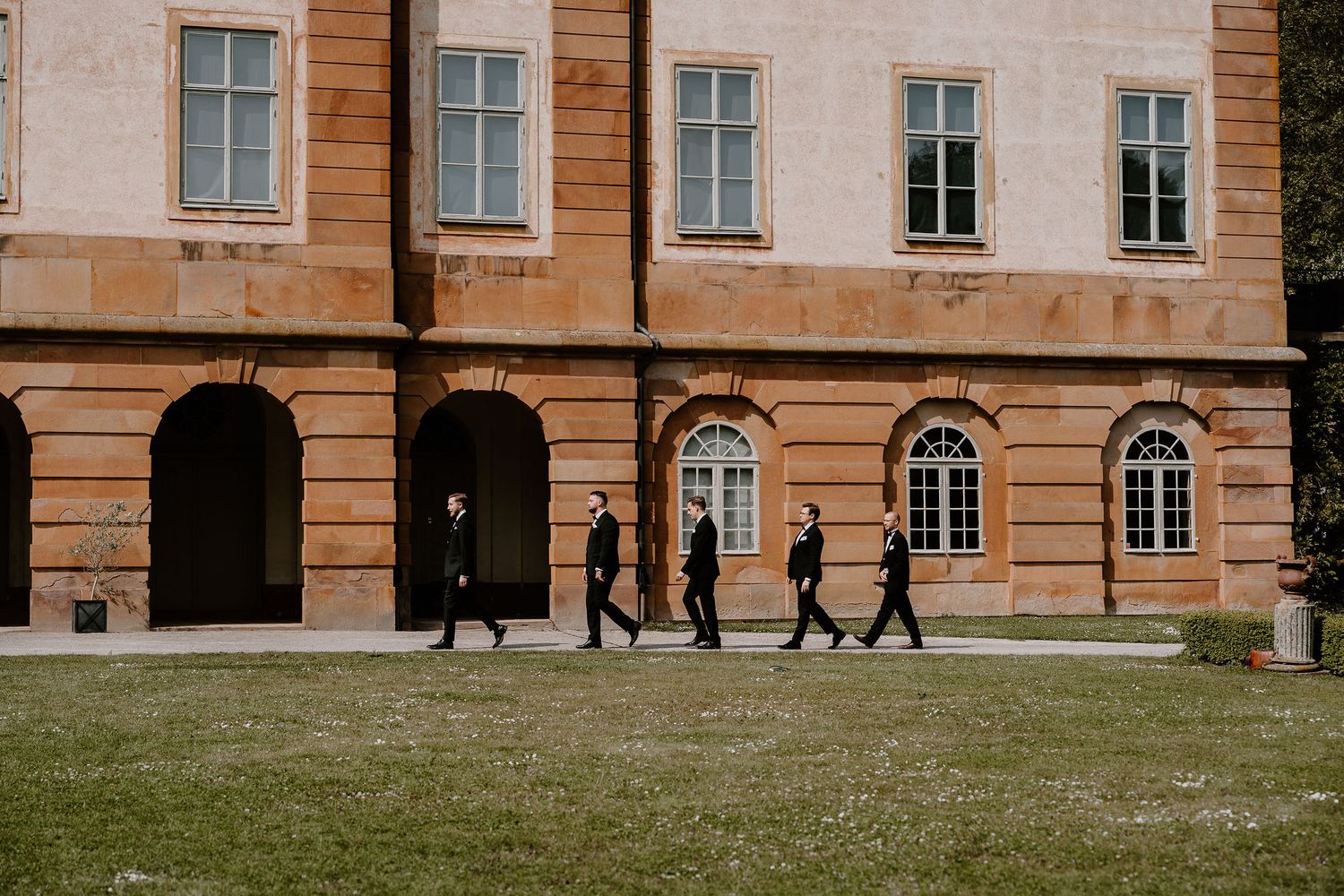 Group of silhouettes walking along a historic building facade with arched windows and pillars on a grassy lawn.