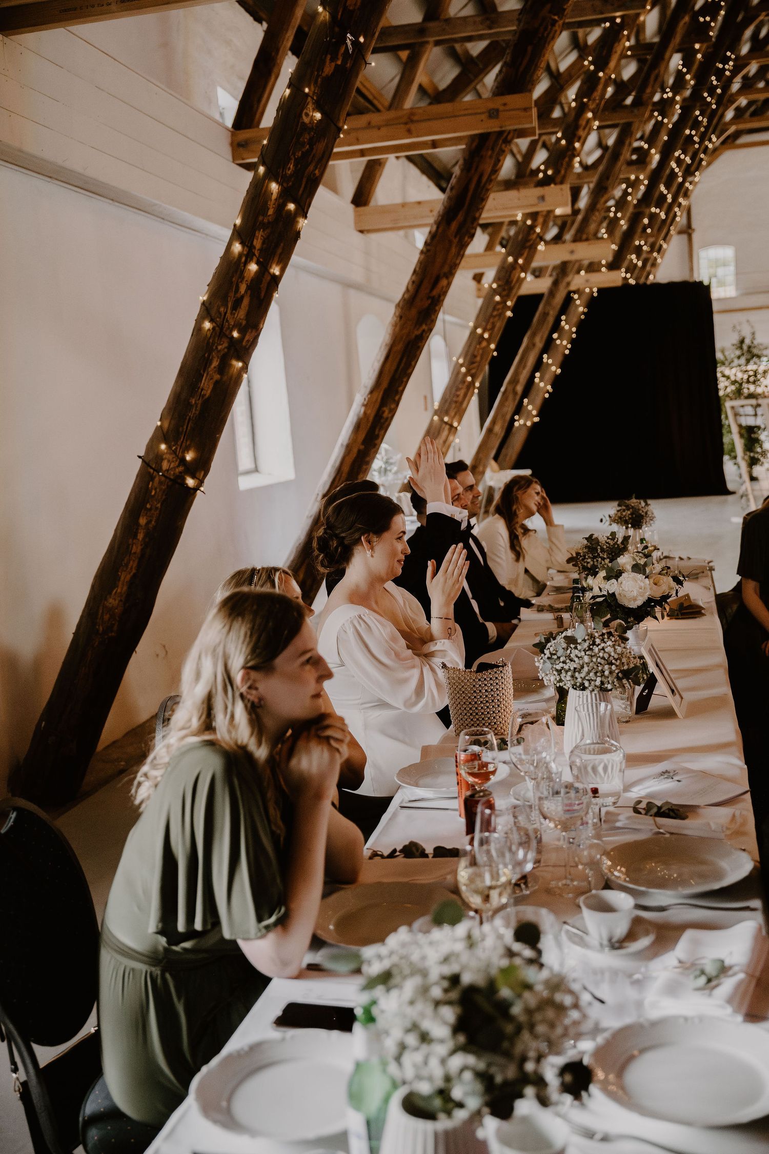 People gather at an elegant table setting under rustic wooden beams decorated with string lights for an intimate celebration.