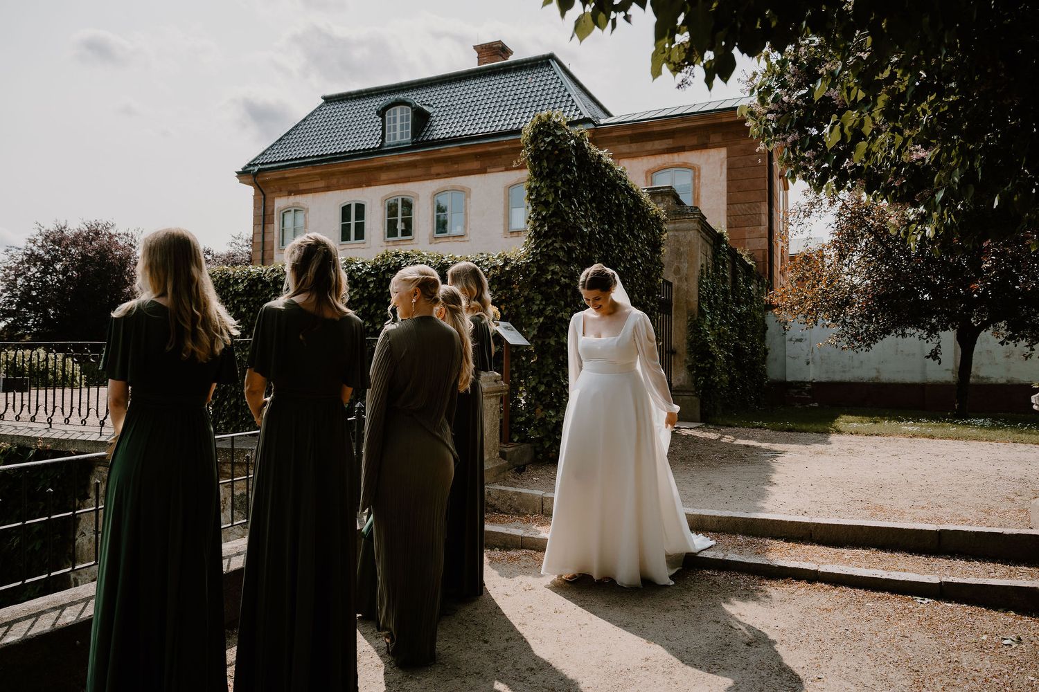 A wedding party in dark formal attire gathers on a sunlit path in front of a stately historic mansion.