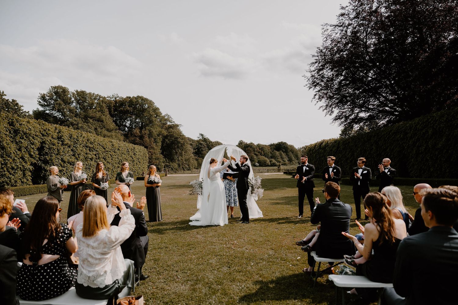 Wedding guests gather outdoors in a scenic field for a rustic countryside celebration near trees at sunset.