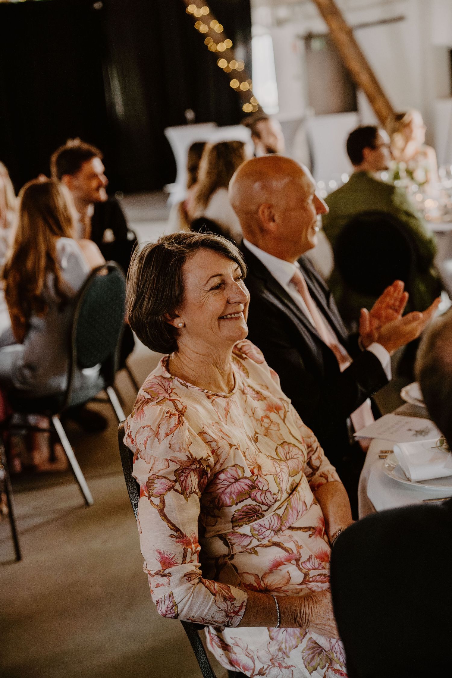 Person in floral dress smiling and laughing during an indoor celebration with string lights in the background.