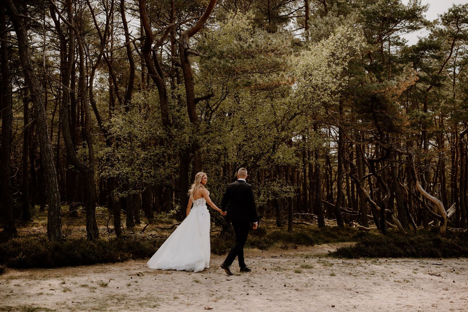 A couple in wedding attire walks hand in hand through a moody forest landscape beside sandy ground.