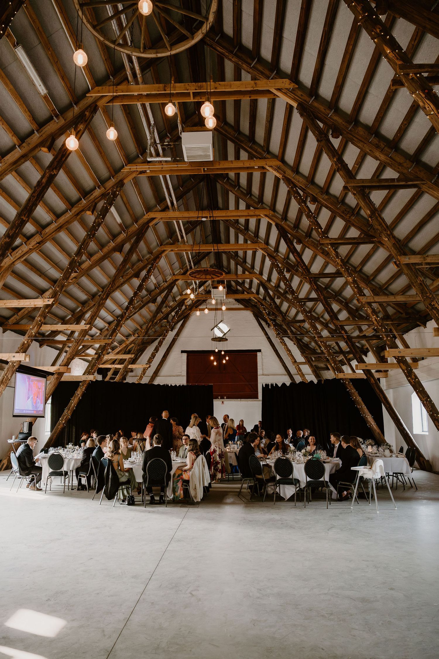 Interior of rustic barn venue with exposed wooden beams and pendant lights during a reception event with guests at round tables.