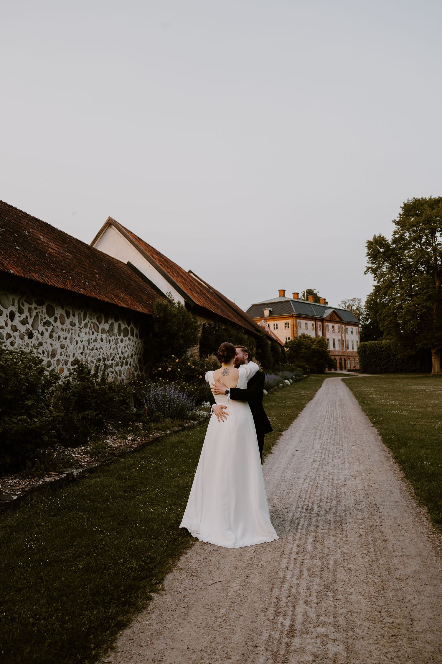 A couple embraces on a gravel path leading to a grand estate with stone walls and garden scenery at dusk.
