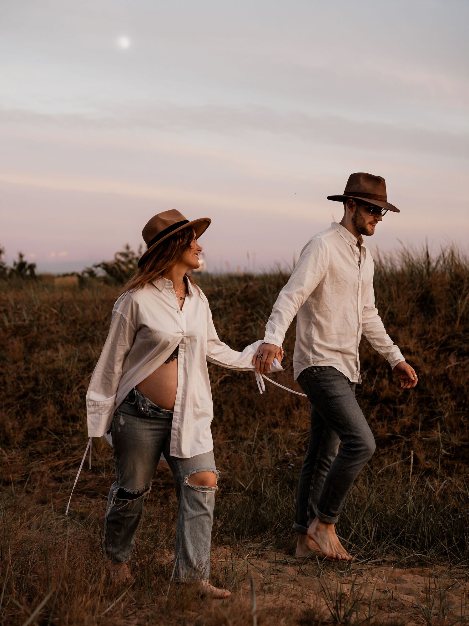 Two people in denim and white shirts with brown hats walking together through a dusty field at sunset.
