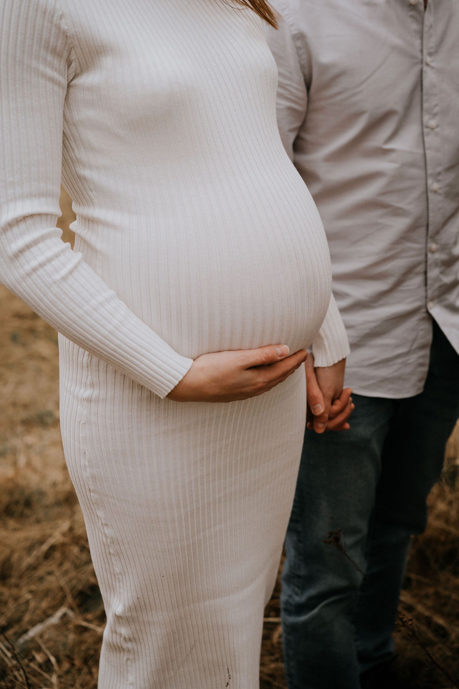 A pregnant belly in a white ribbed sweater dress is gently held while standing next to someone wearing denim.
