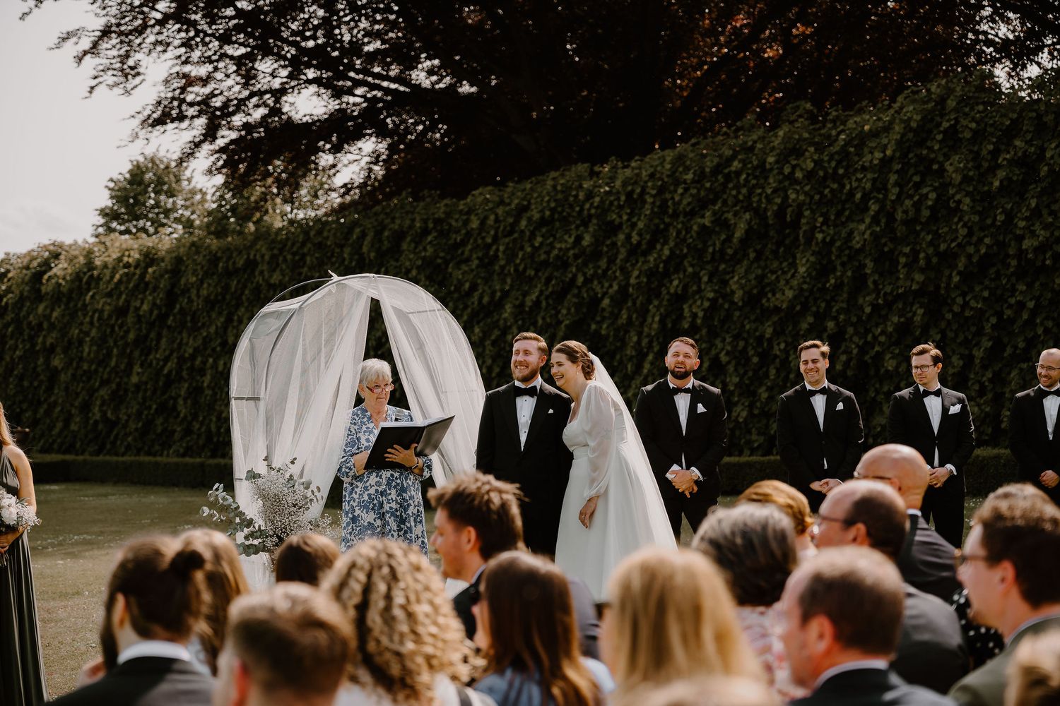 An elegant outdoor wedding ceremony with flowing white drapery and tall trees providing a natural backdrop.