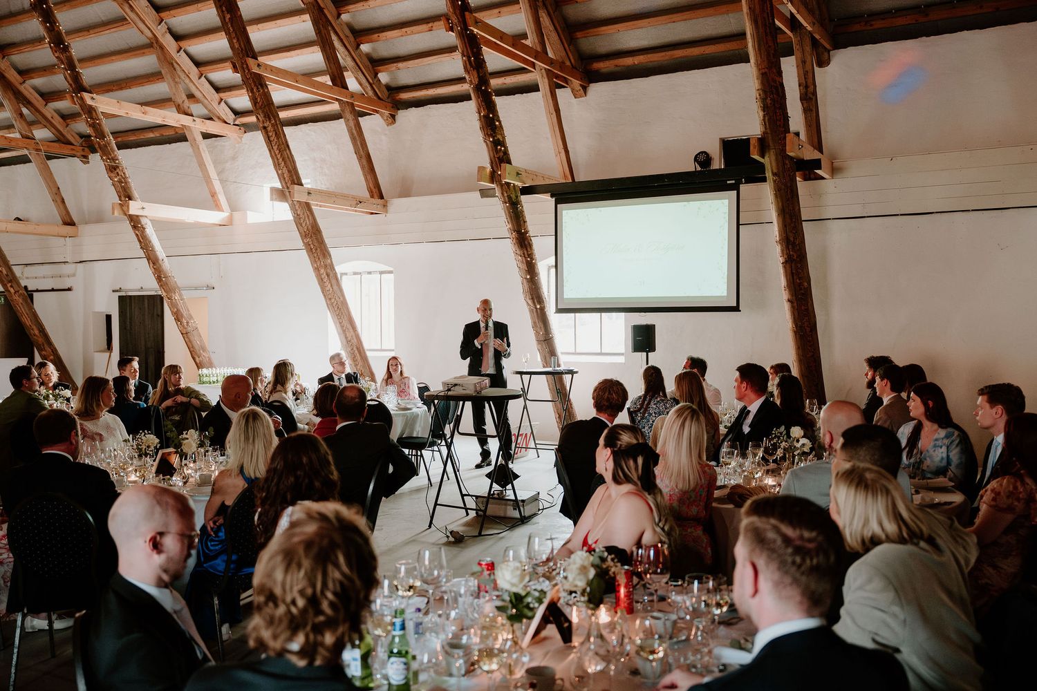 Wedding reception guests seated at tables watch a presentation on projector screen in rustic venue with exposed beams.