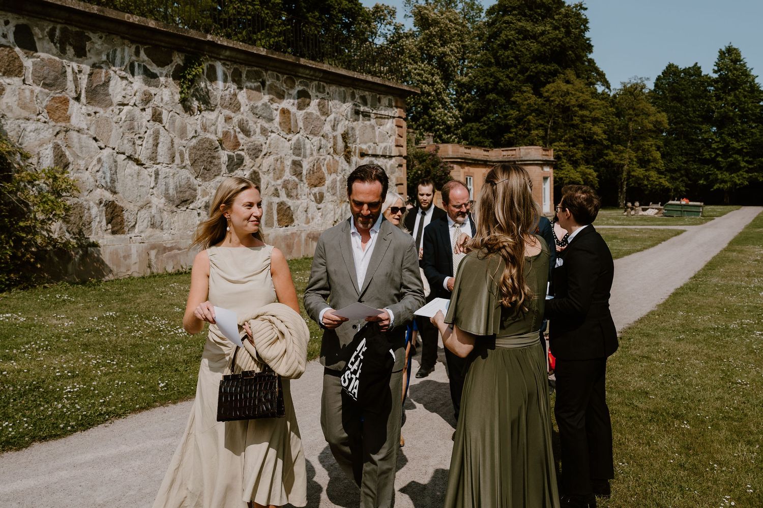 Small outdoor wedding ceremony with attendees gathered near rustic stone wall and green lawn.