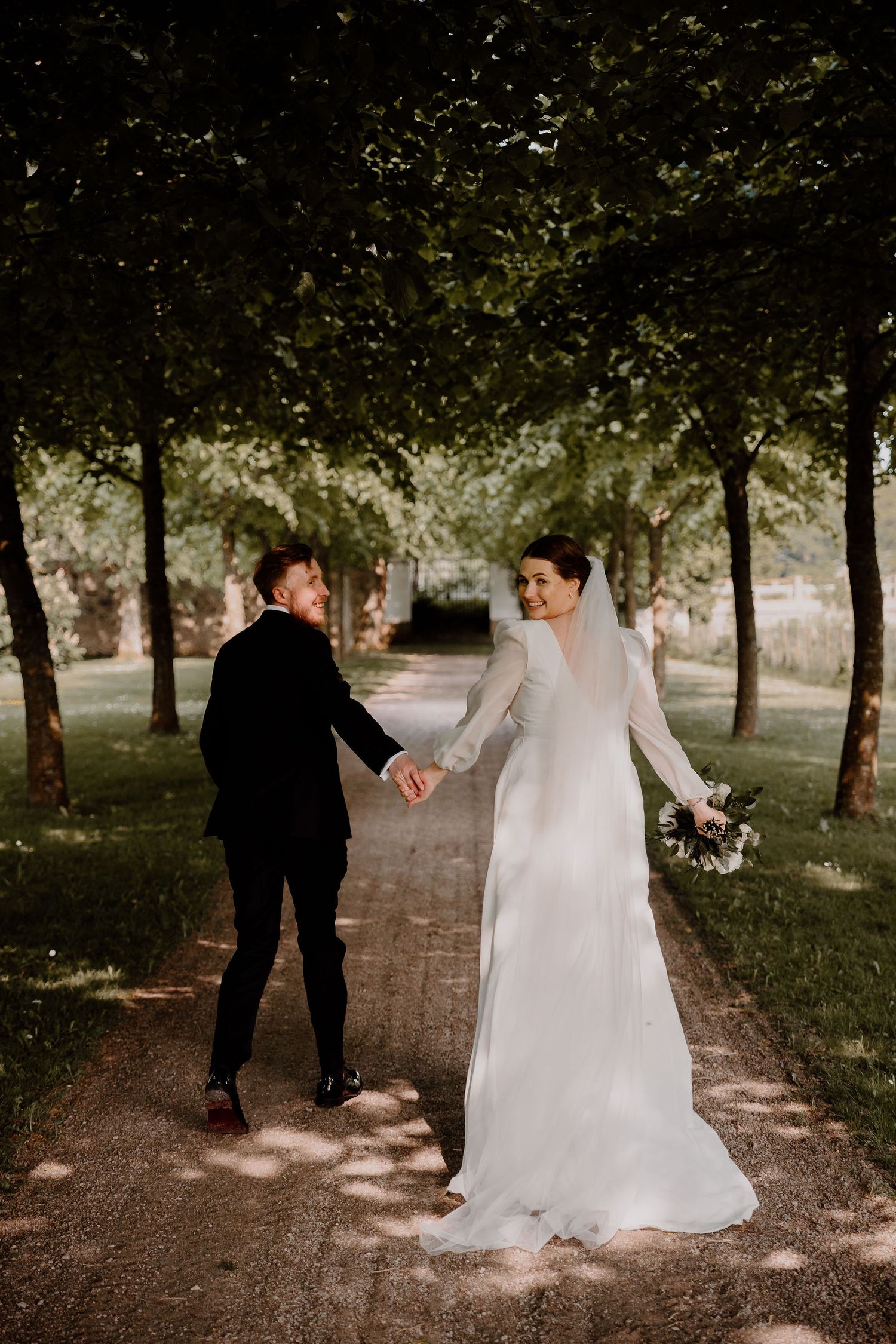Newlywed couple walks hand in hand along a tree-lined path in the afternoon sunlight.
