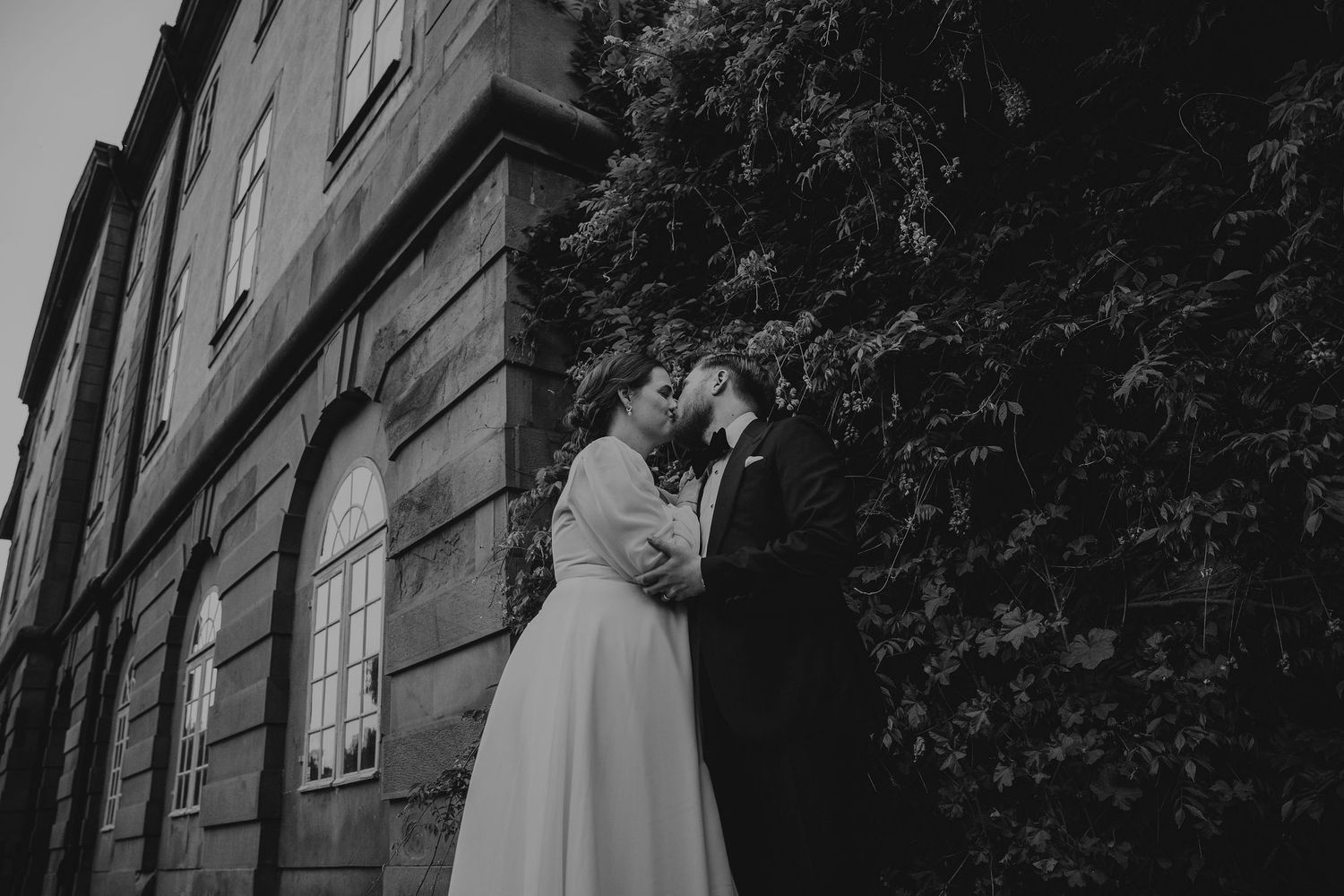A black and white wedding photograph of a couple sneaking a kiss outside a historical brick building.