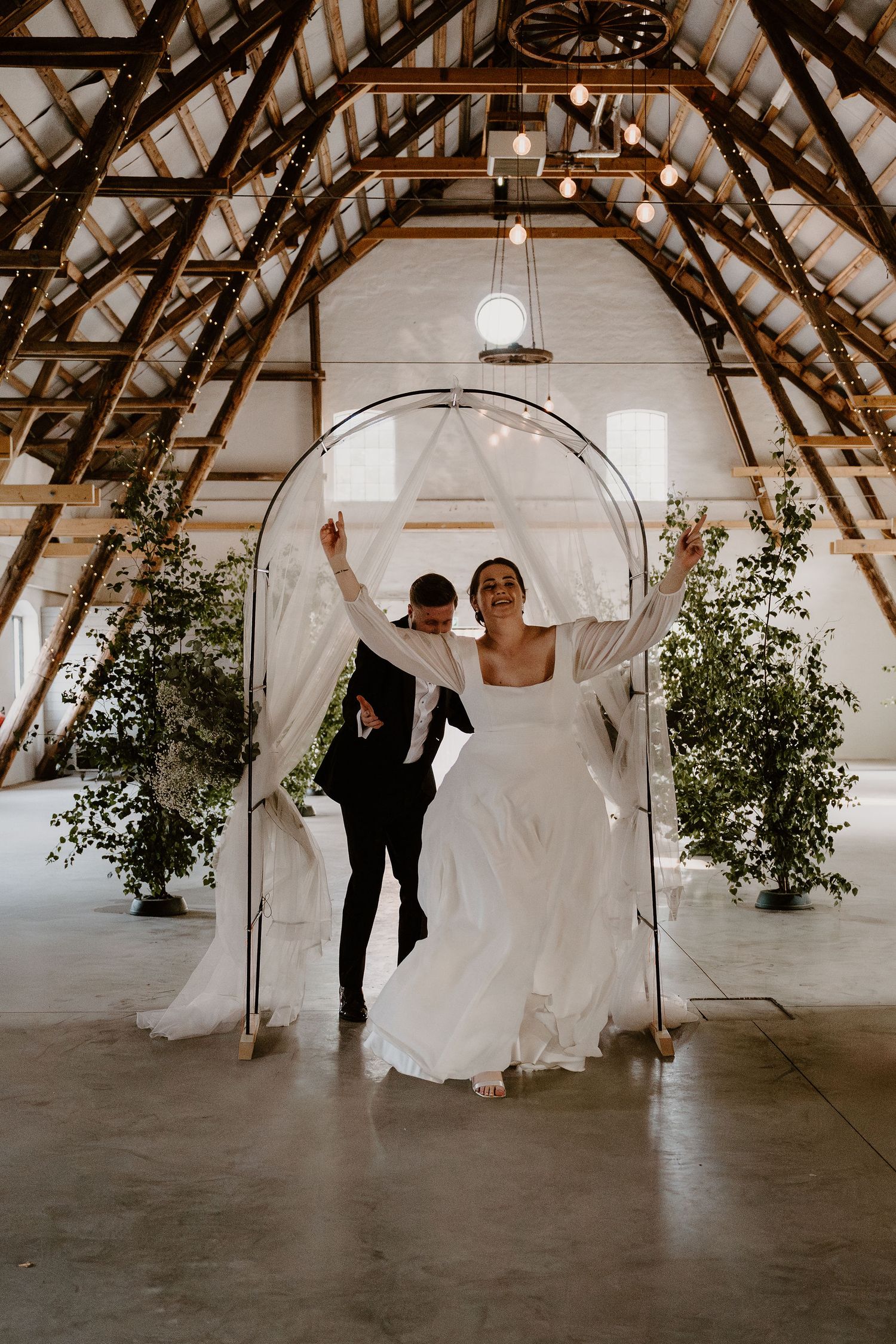 A couple celebrates under a rustic wooden beam ceiling with greenery and a decorative arch in an elegant barn venue.
