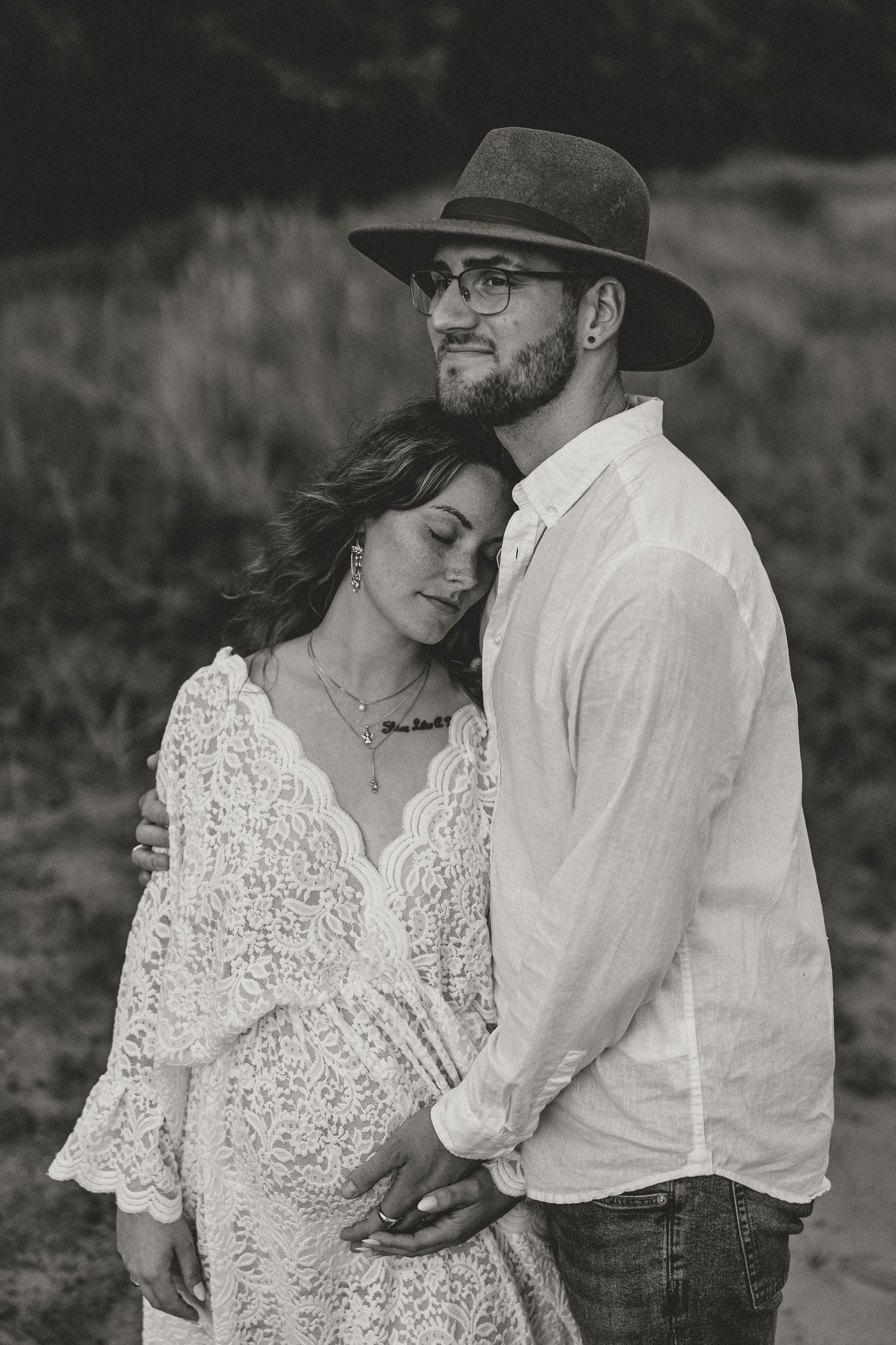 Black and white portrait of an expecting couple in intimate embrace, with woman in lace dress and man wearing hat and glasses.
