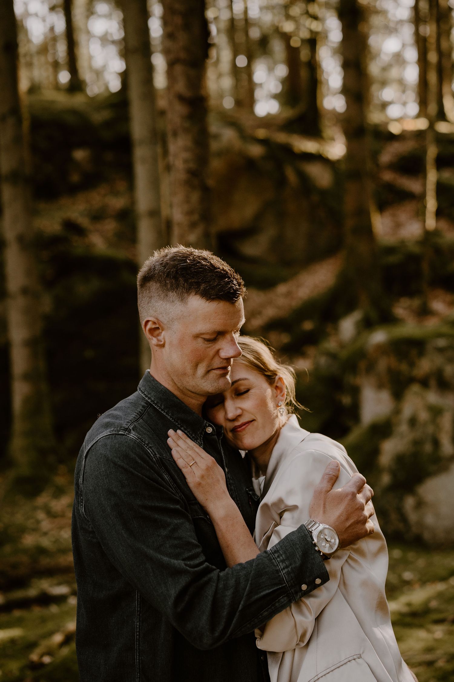 A tender romantic moment captured between two people embracing in warm golden sunlight filtering through a forest.