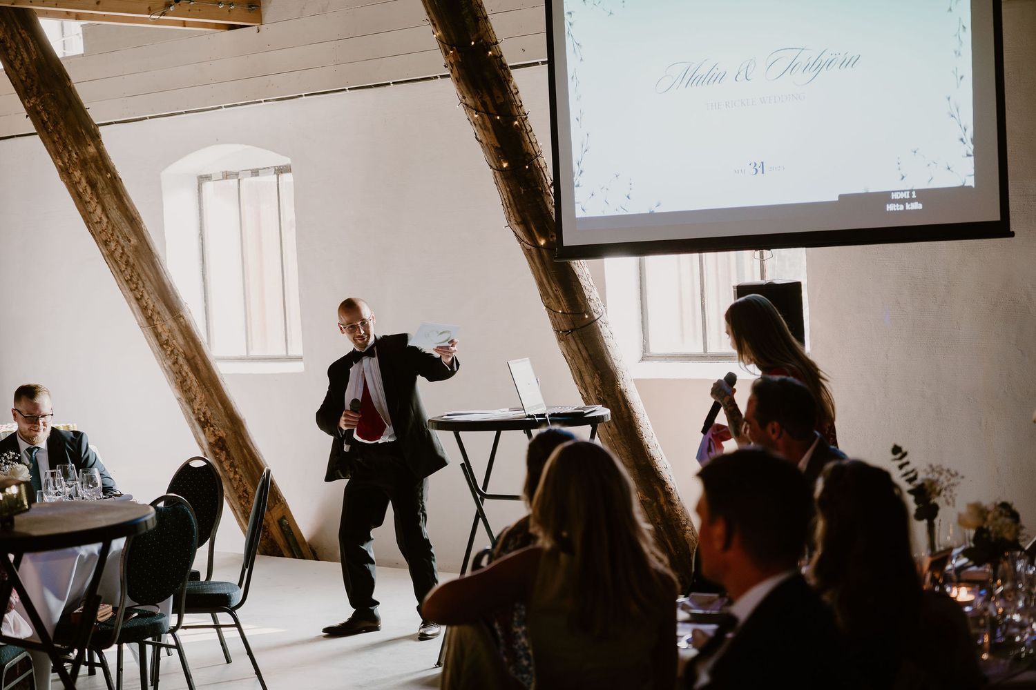 People gather in a rustic venue with exposed wooden beams during a dynamic presentation with a projector screen.