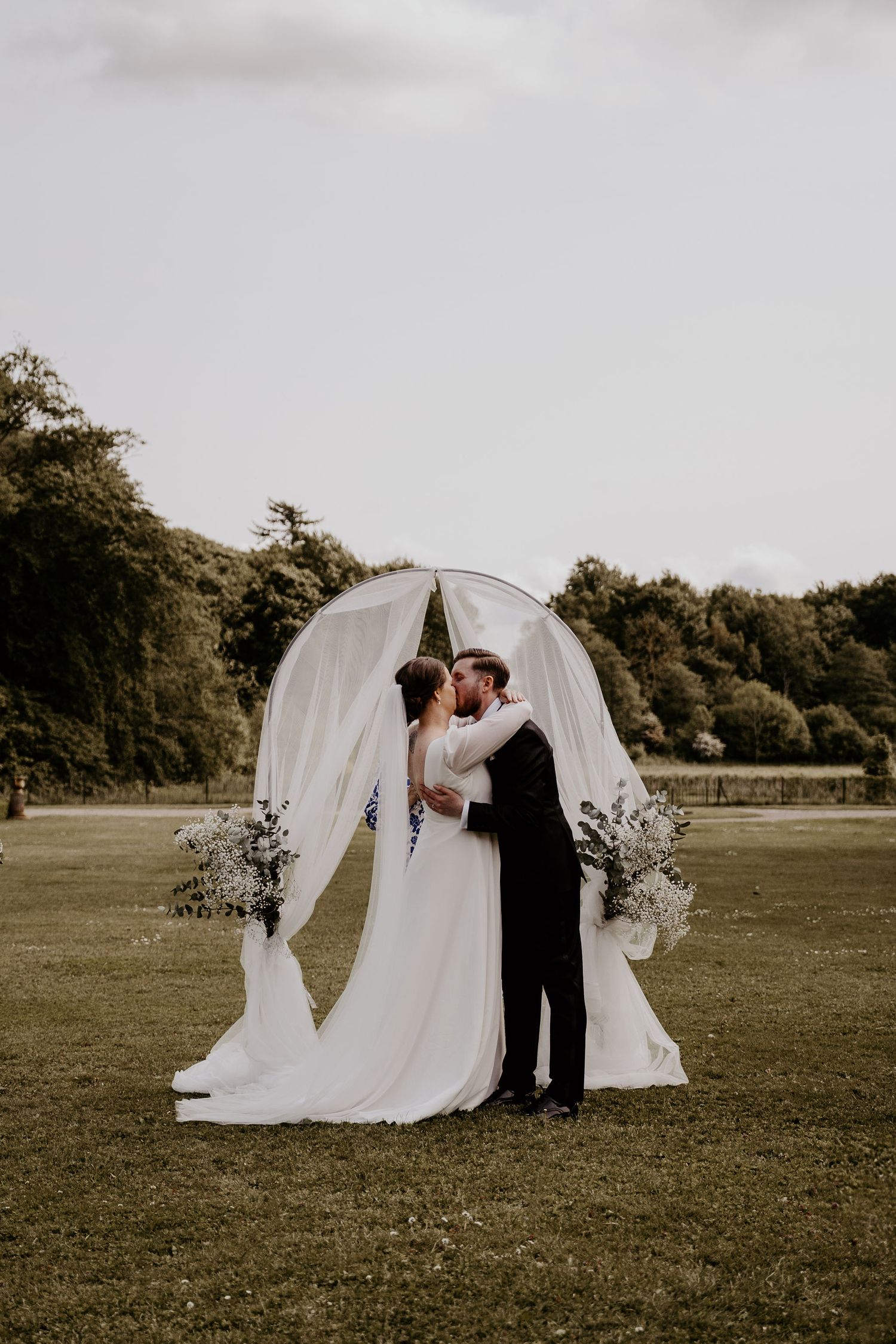 Five aesthetic vintage-style wedding ceremony photos of a couple under a white draped arch in an open field display romantic moments.