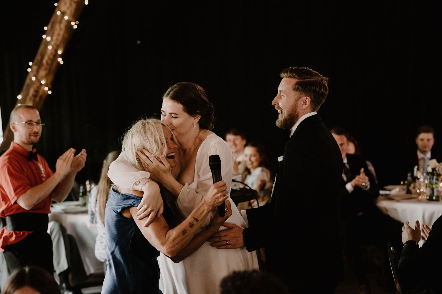A crowd of celebratory wedding guests at a reception with dark moody lighting throw their hands up in excitement.