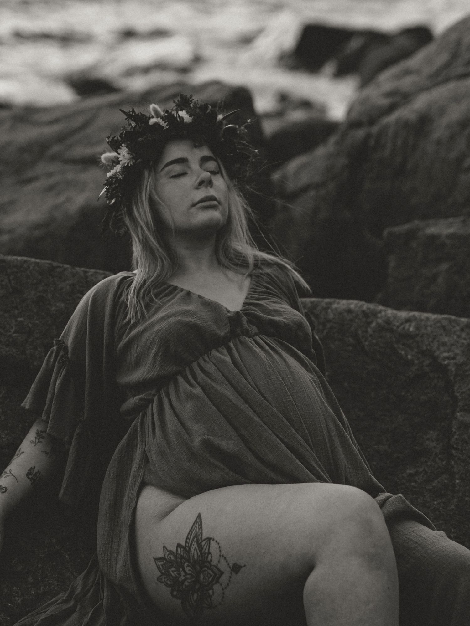 Black and white artistic portrait of figure in floral crown with visible tattoo posed among rocks in ethereal style.