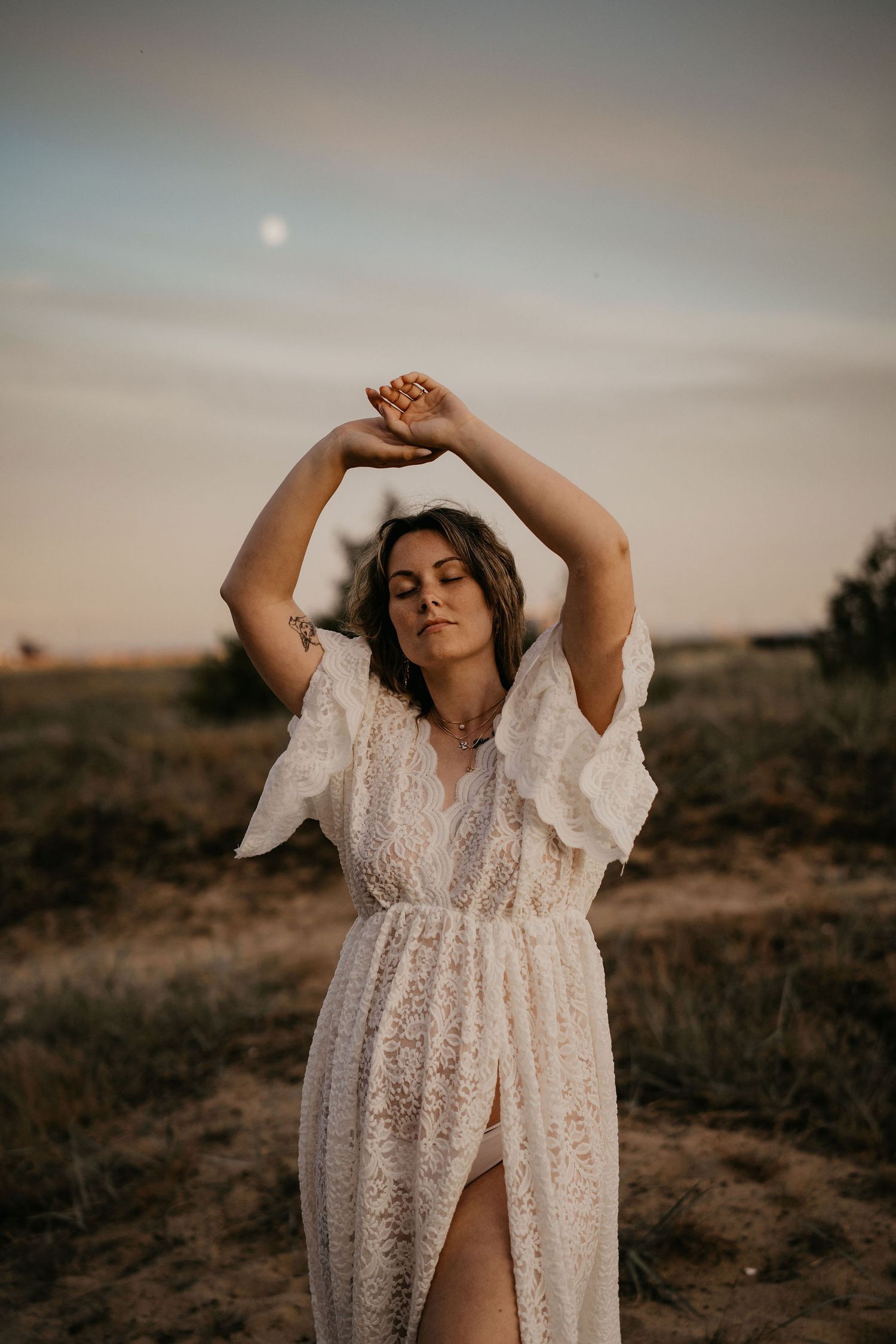 Person in a flowing white lace dress poses elegantly with raised arms in a sunset field.