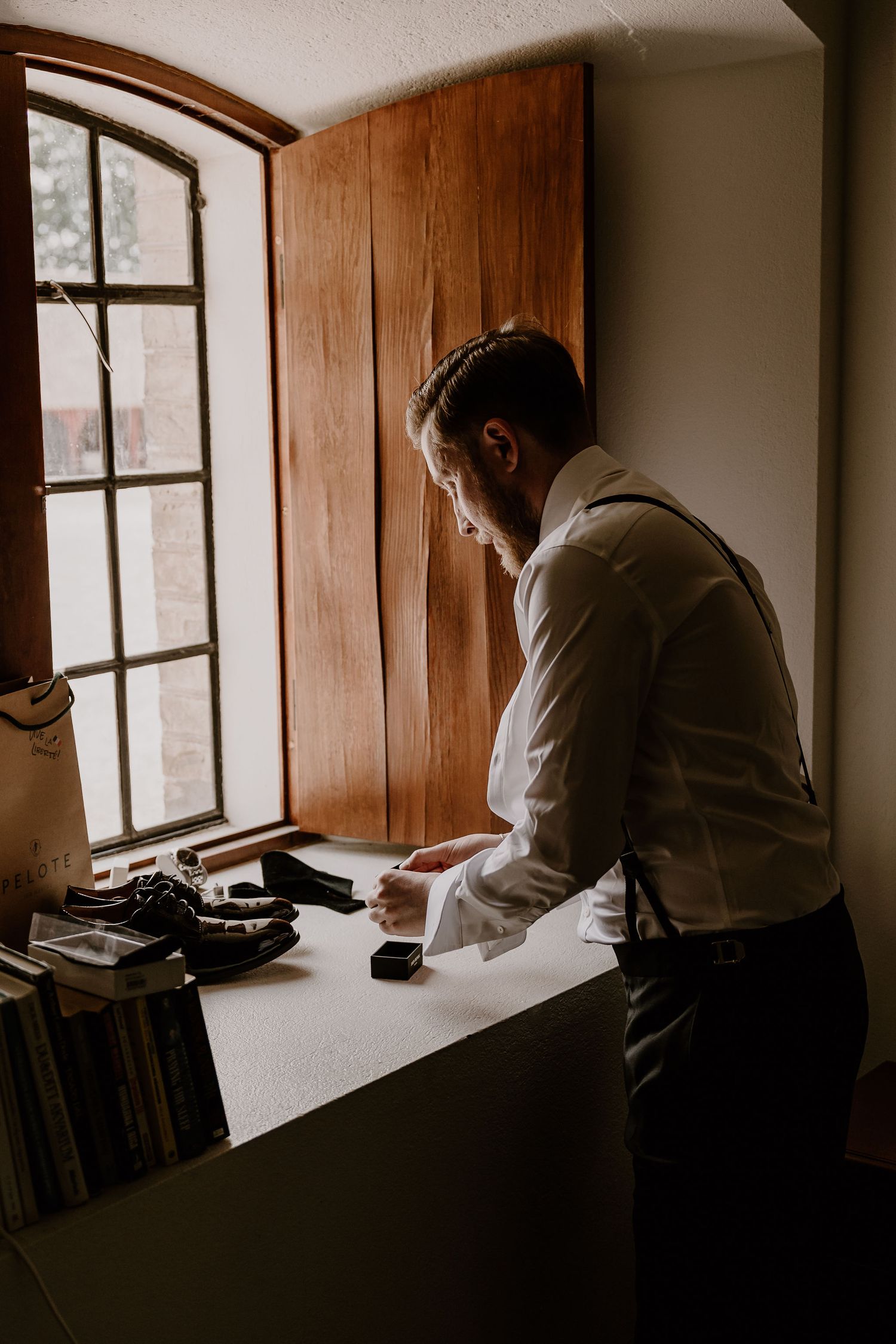 Person in white dress shirt and suspenders stands at window sill next to vintage typewriter in moody lighting.