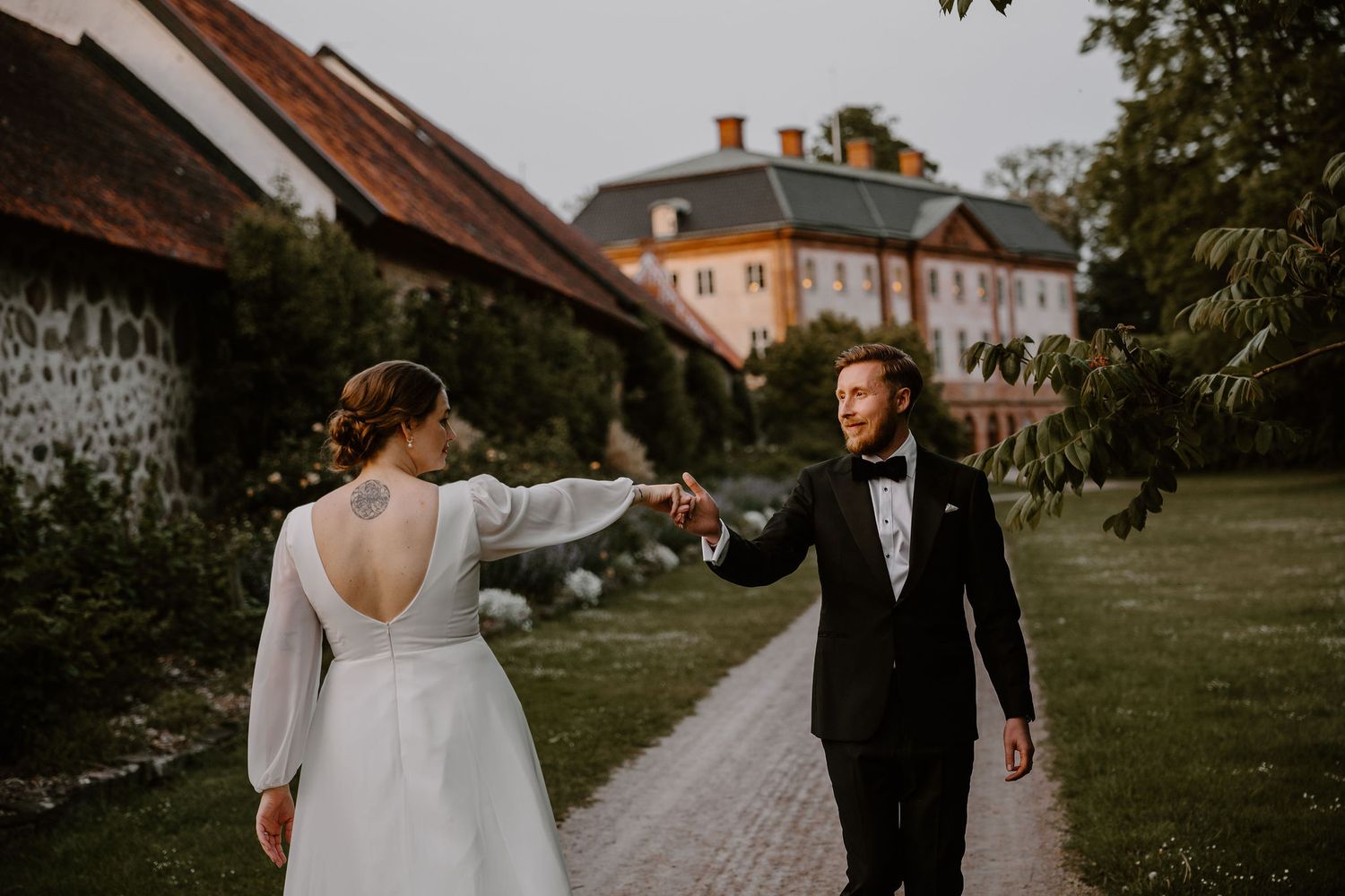 A wedding couple holding hands and dancing on a garden path near a historic manor house.