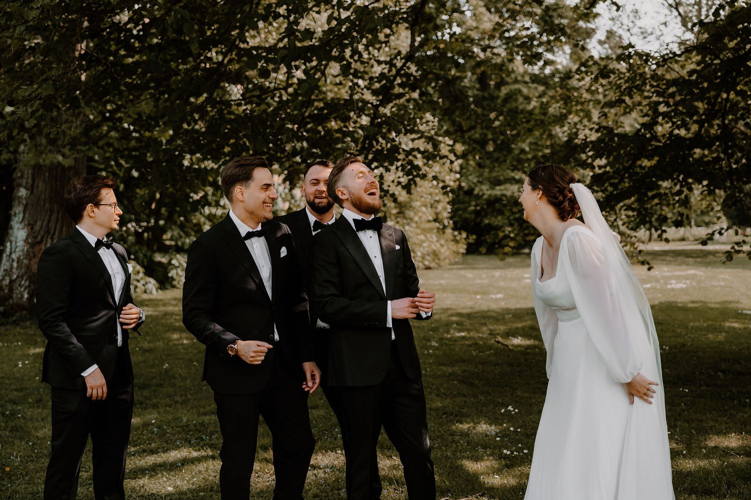 A wedding party shares a candid moment outdoors under trees during a ceremony in natural sunlight.