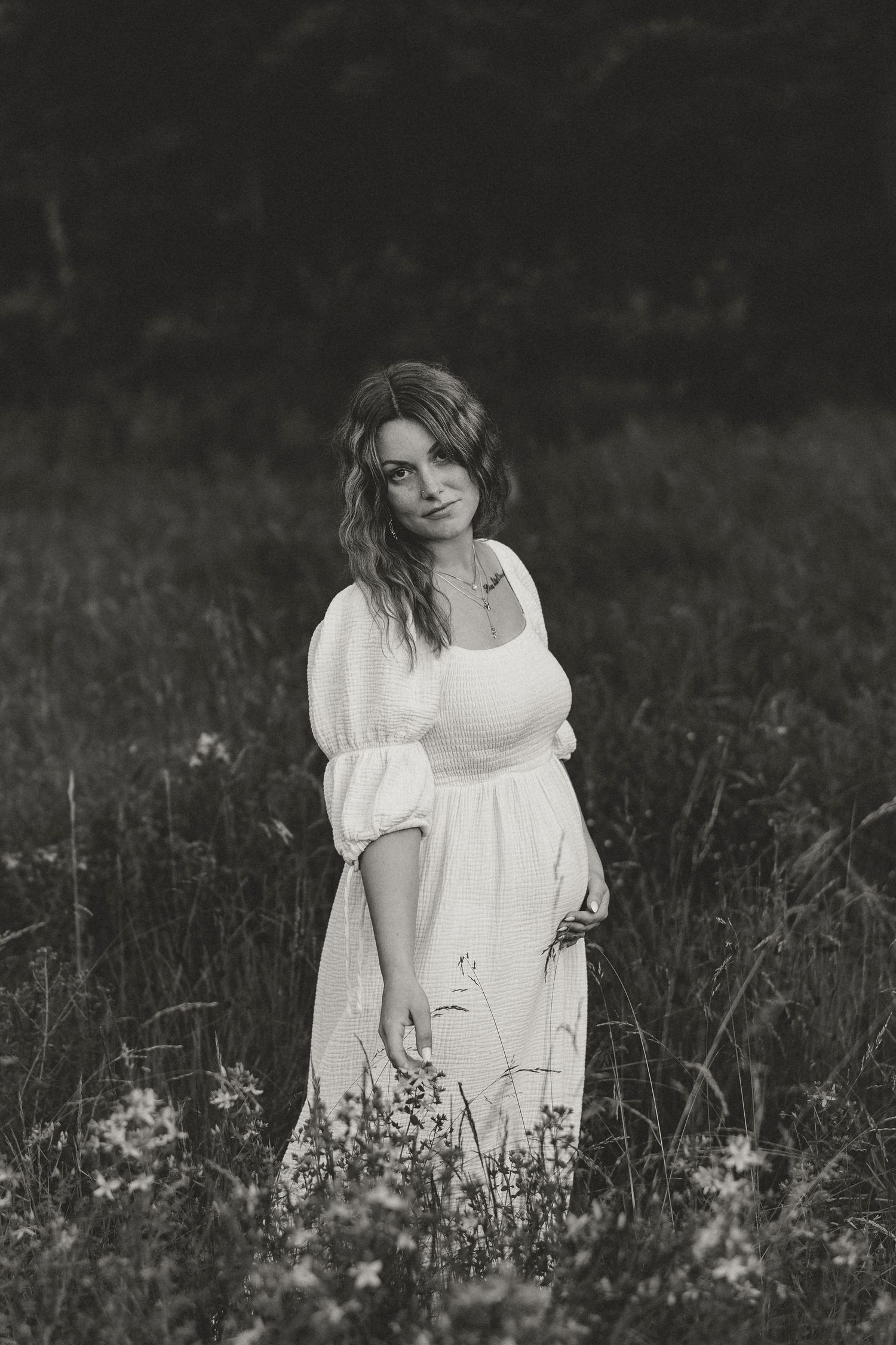 Black and white portrait of someone in a white dress standing in a meadow with wildflowers.