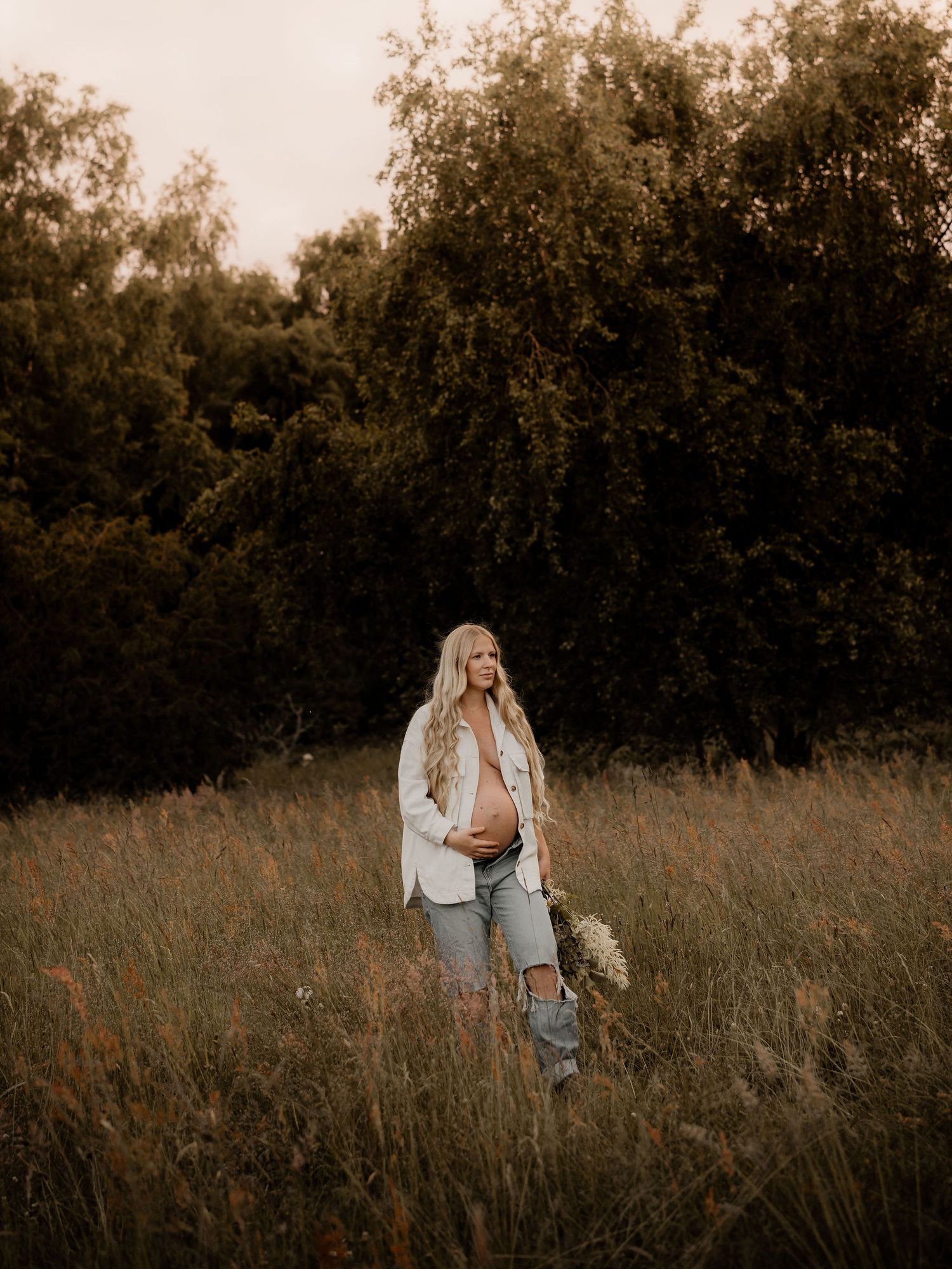 Person in casual attire poses in a rustic field during sunset maternity photo session.