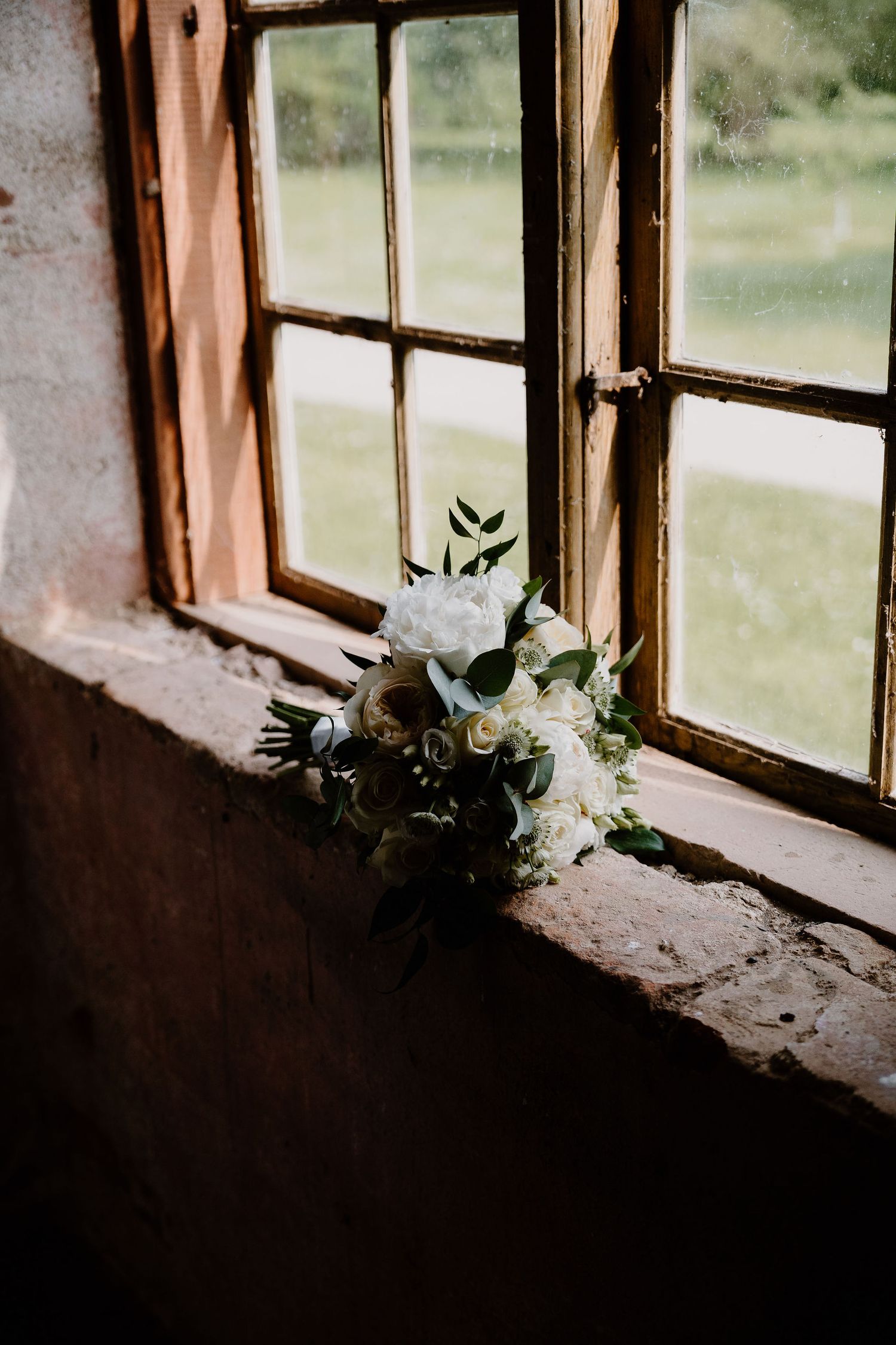 A white floral bouquet sits on a rustic windowsill in moody natural lighting with vintage wooden window frames.