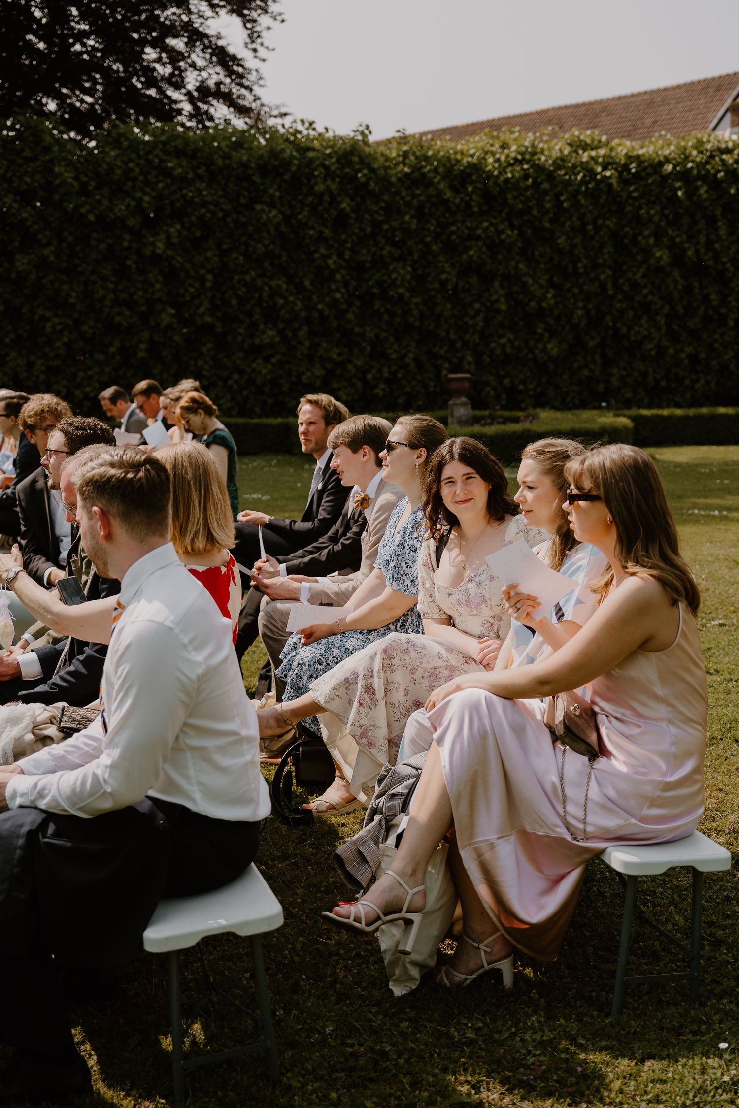 Wedding guests seated in chairs on a lawn during an outdoor ceremony in warm sunlight with a lush hedge backdrop.