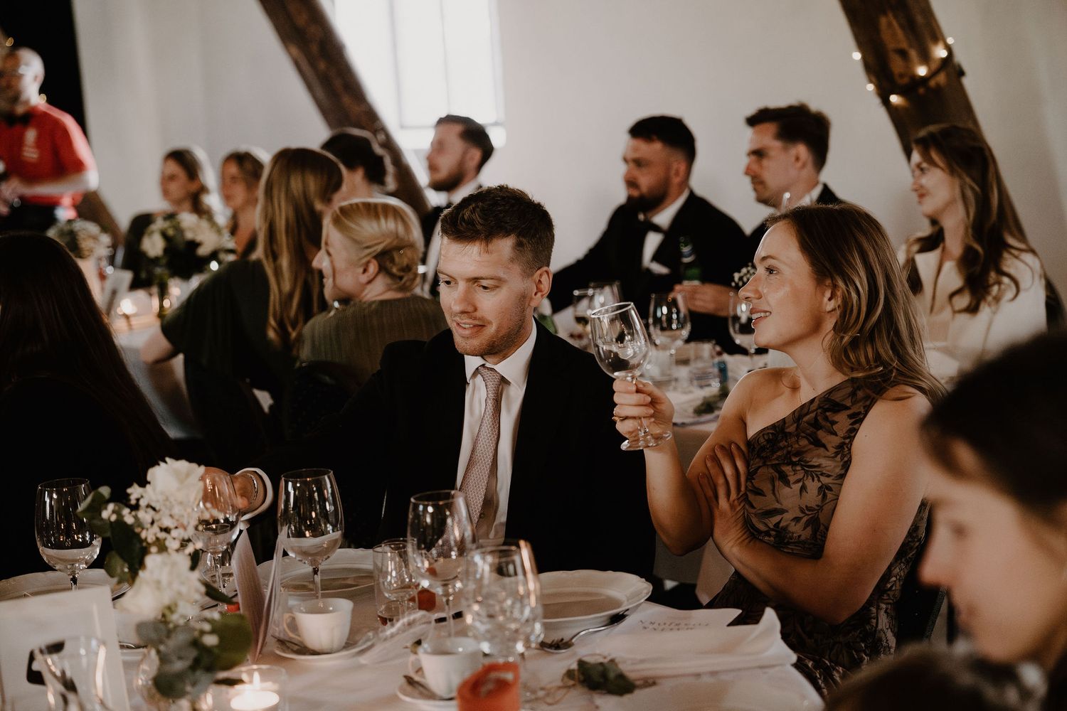 Wedding guests socialize and laugh around elegantly set dining tables at an indoor reception venue.