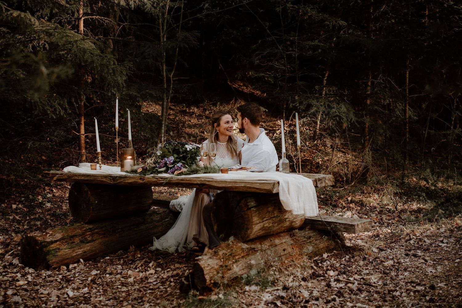 Moody autumn wedding scene with two people sitting at rustic wooden table decorated with candles in forest setting.