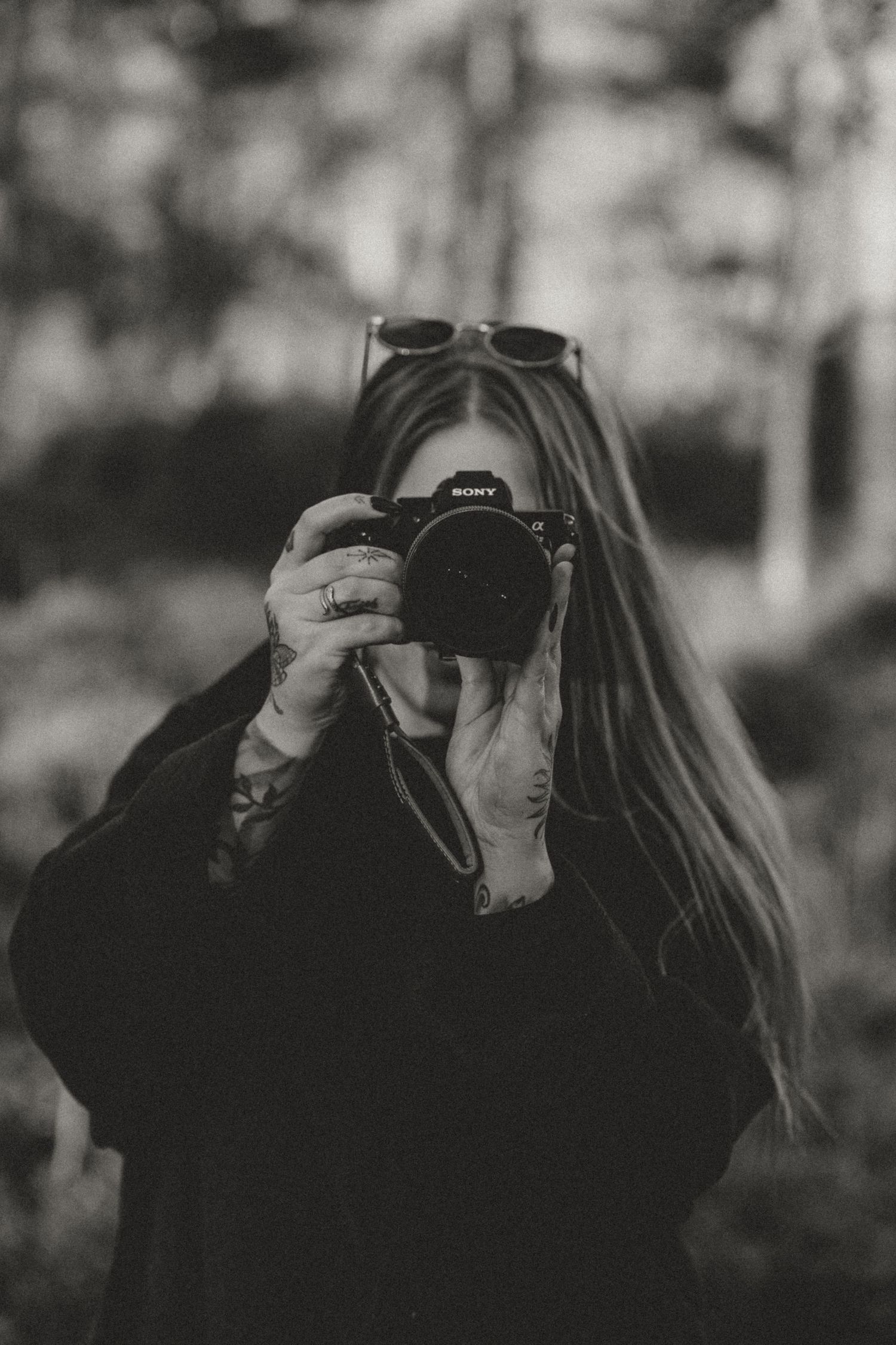 Black and white portrait of a photographer with long hair holding a professional camera against a blurred background.