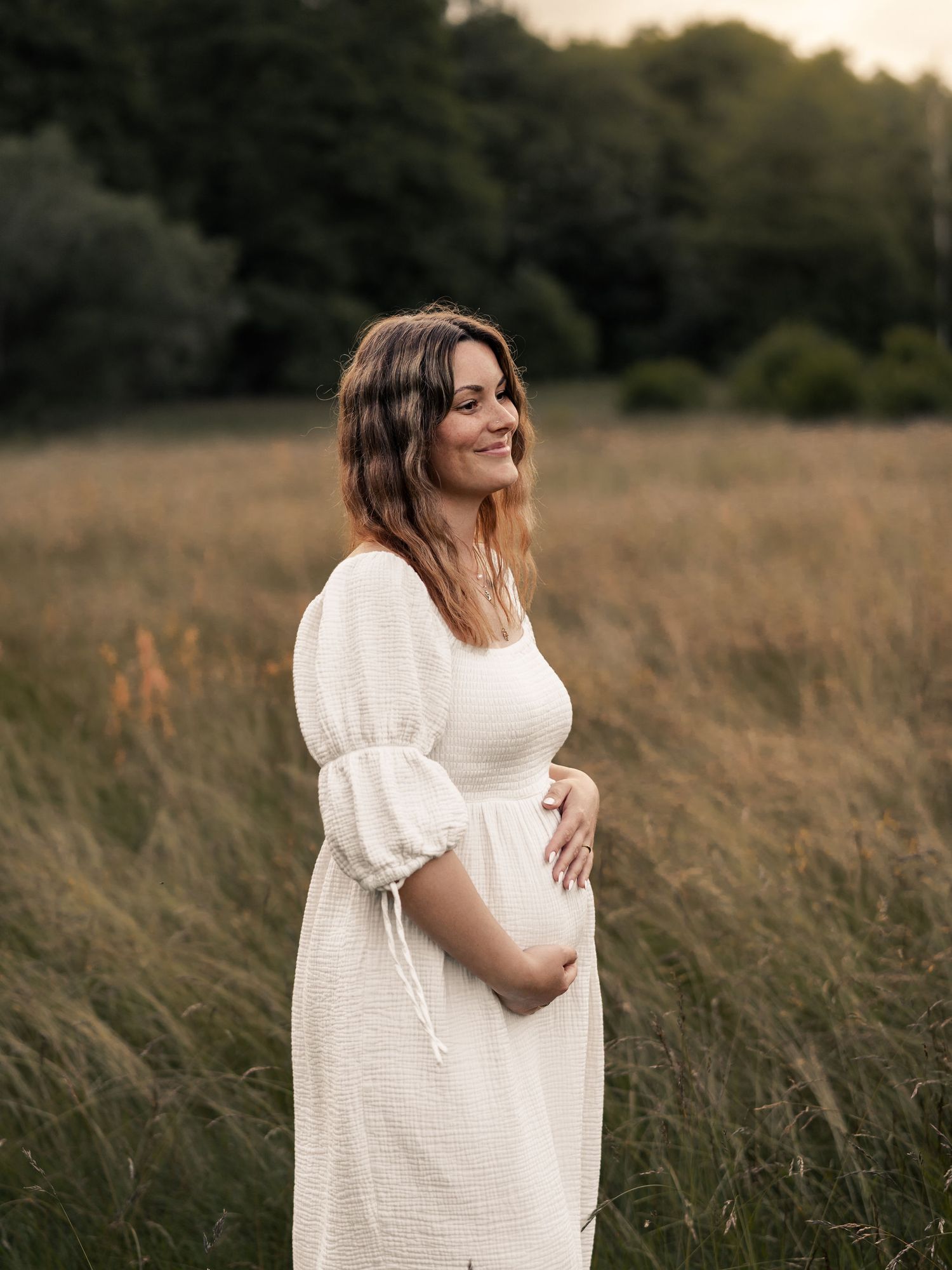 Expectant woman in white dress smiles while standing in tall grass during golden hour.