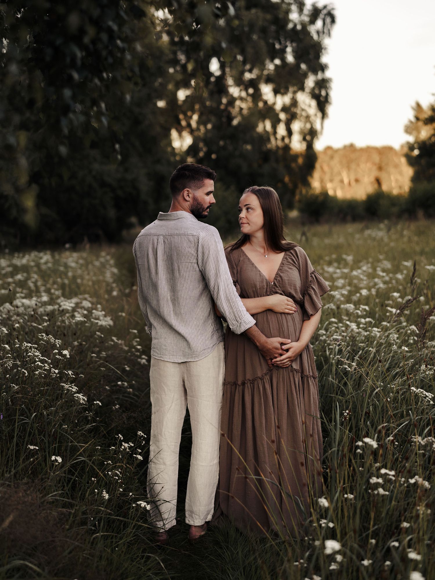 A couple stands together in a wildflower meadow at sunset, with the woman wearing a flowing brown dress and cradling her belly.
