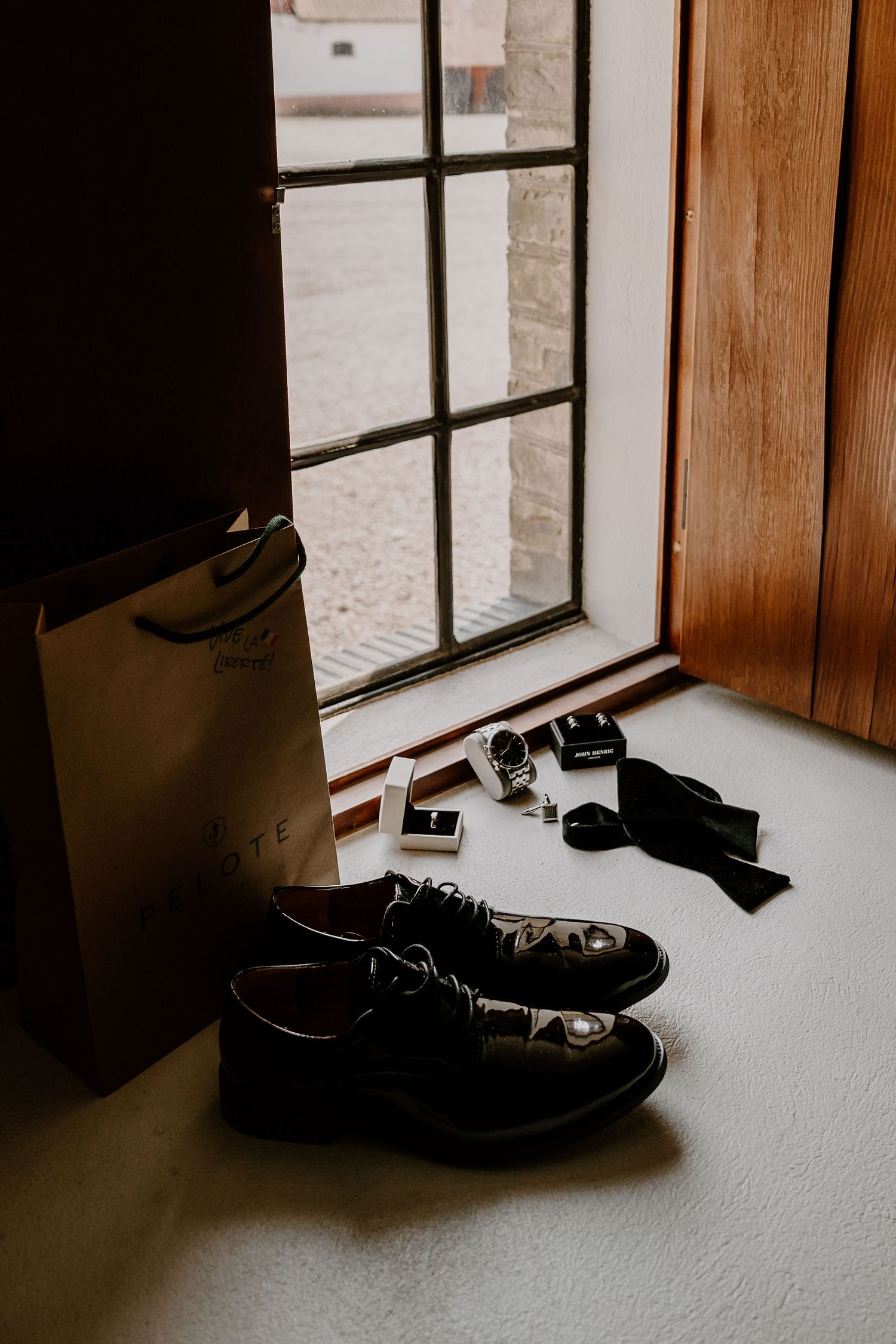 A pair of black dress shoes sits by a windowsill next to a paper bag and some scattered small objects.
