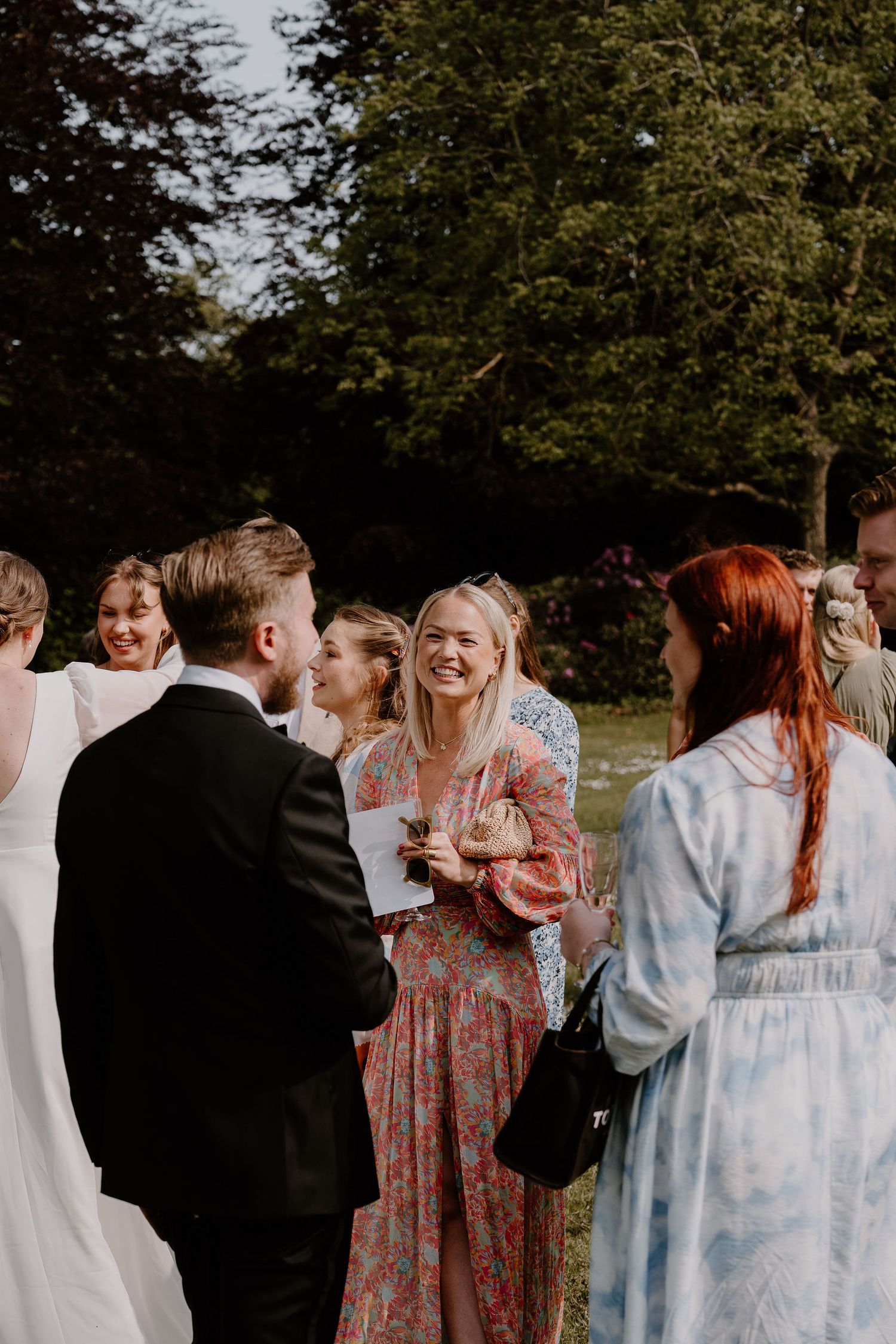 Wedding guests gather outdoors on a sunny day, sharing joyful laughter and conversation at an elegant garden reception.